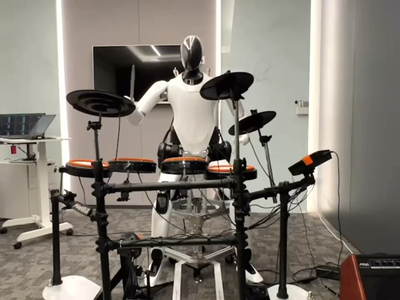 A black and white humanoid robot sits at an electronic drum kit