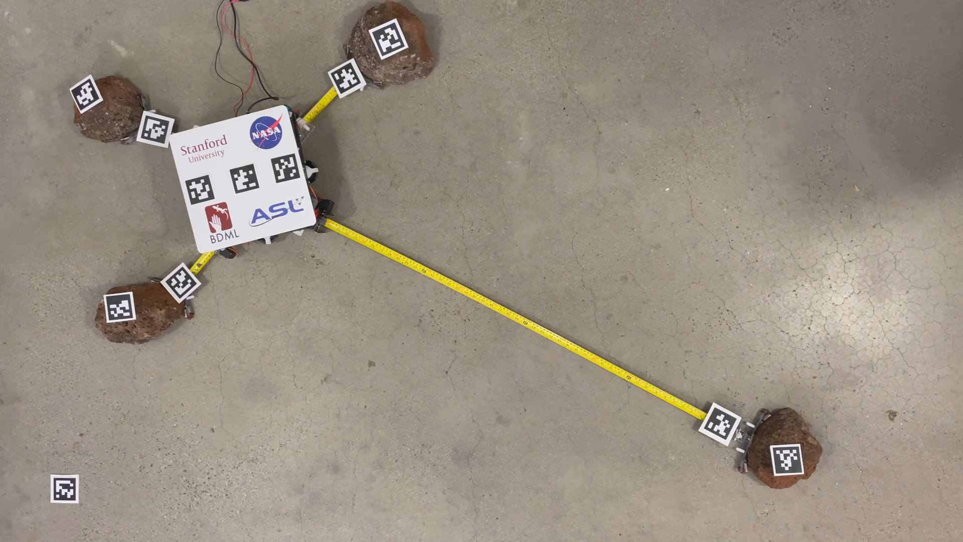 A square white robot on a concrete floor attaches to rocks using grippers on the ends of extended metal measuring tapes.