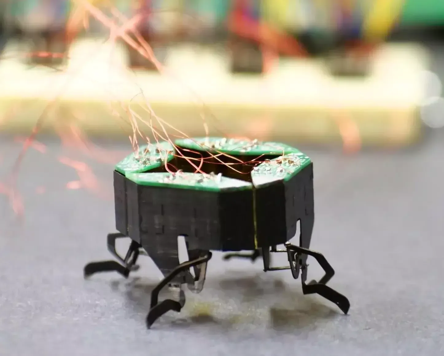 A close up photo of a tiny robot with six black legs and a circuit board on top, attached to a breadboard in the background through copper wires.