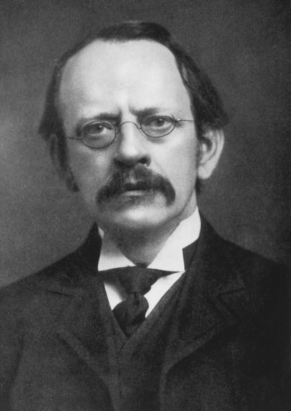 In a black-and-white image of the British physicist J.J. Thomson, he has a dark mustache and wire-rimmed glasses.