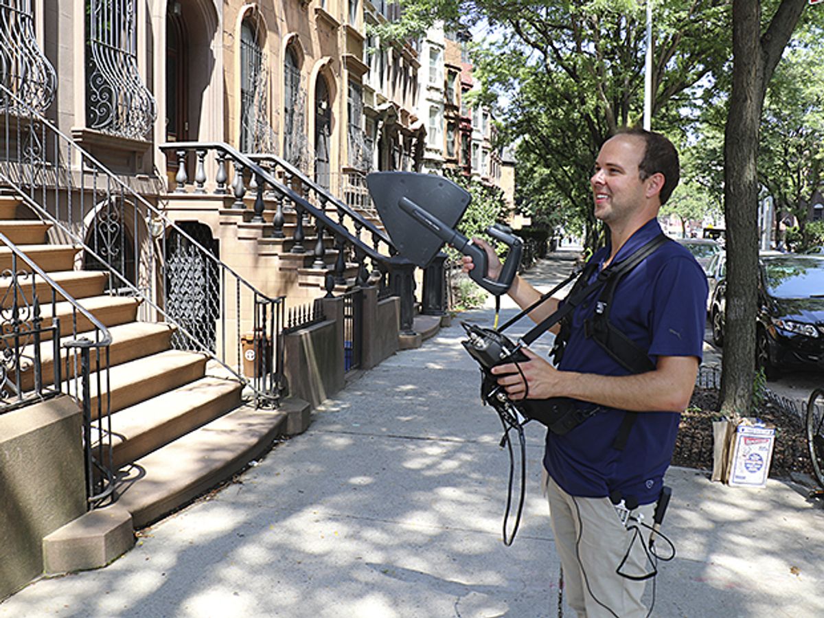 In front of several brownstone buildings, an equipment-laden man holds a gray instrument shaped like a spade.