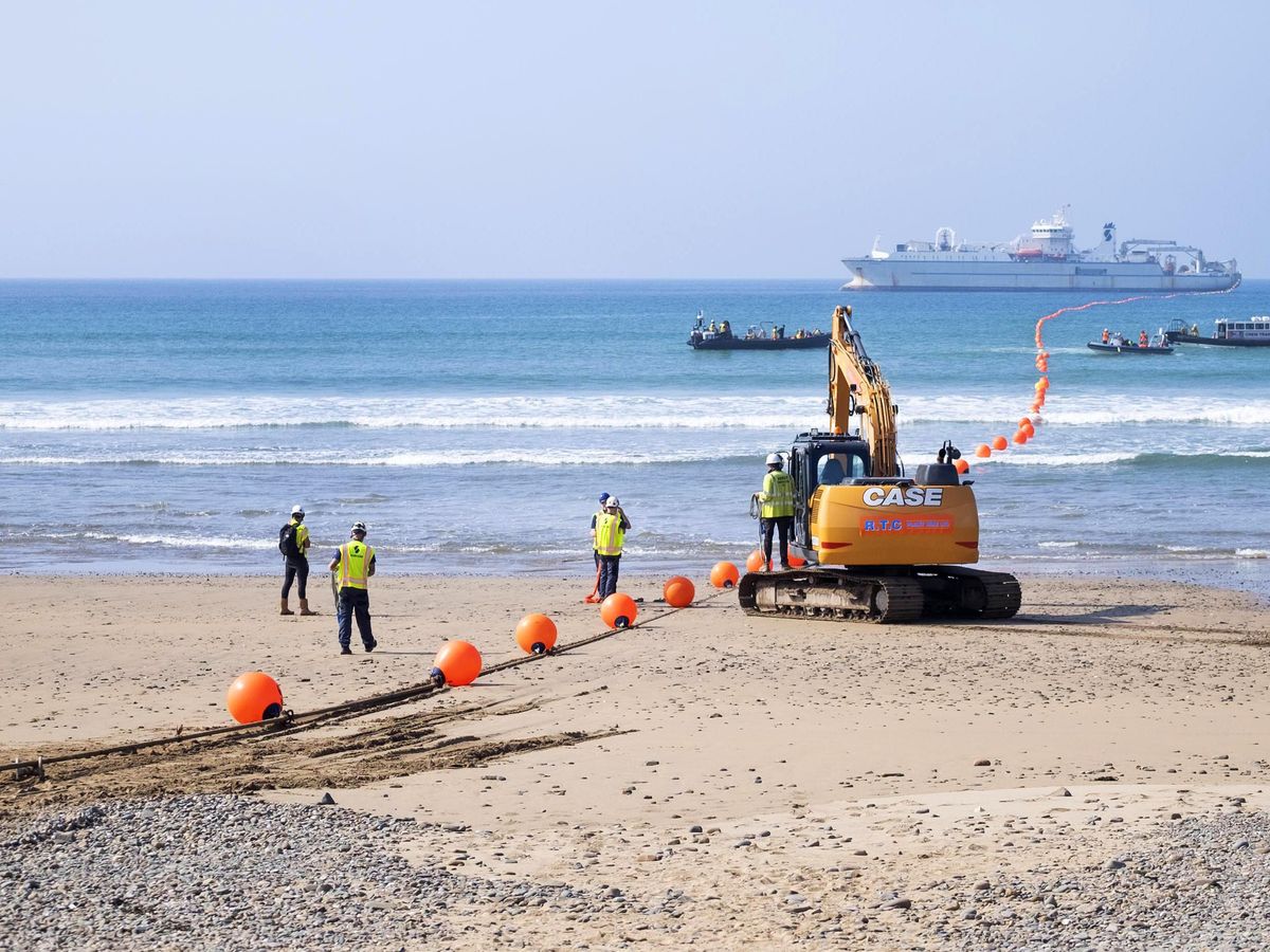 In the background, a large white ship in water. A long cable, covered in orange buoys travels from the ship to the beachy shore, where a digger and 5 people in hard hats and bright vests look on.