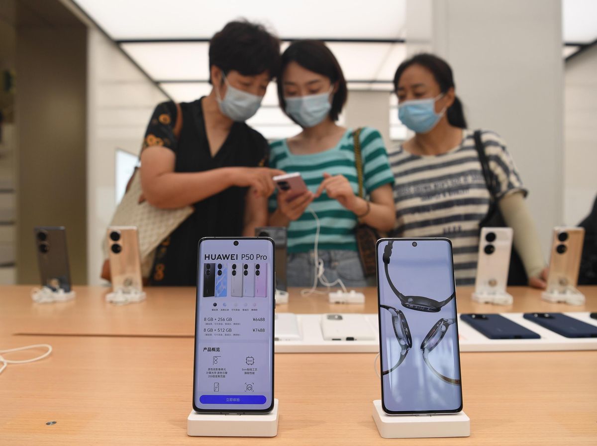 In the foreground, 2 rectangular phones sitting on white docks say Huawei P50 Pro. In the background, 3 women in masks look at phones on display.