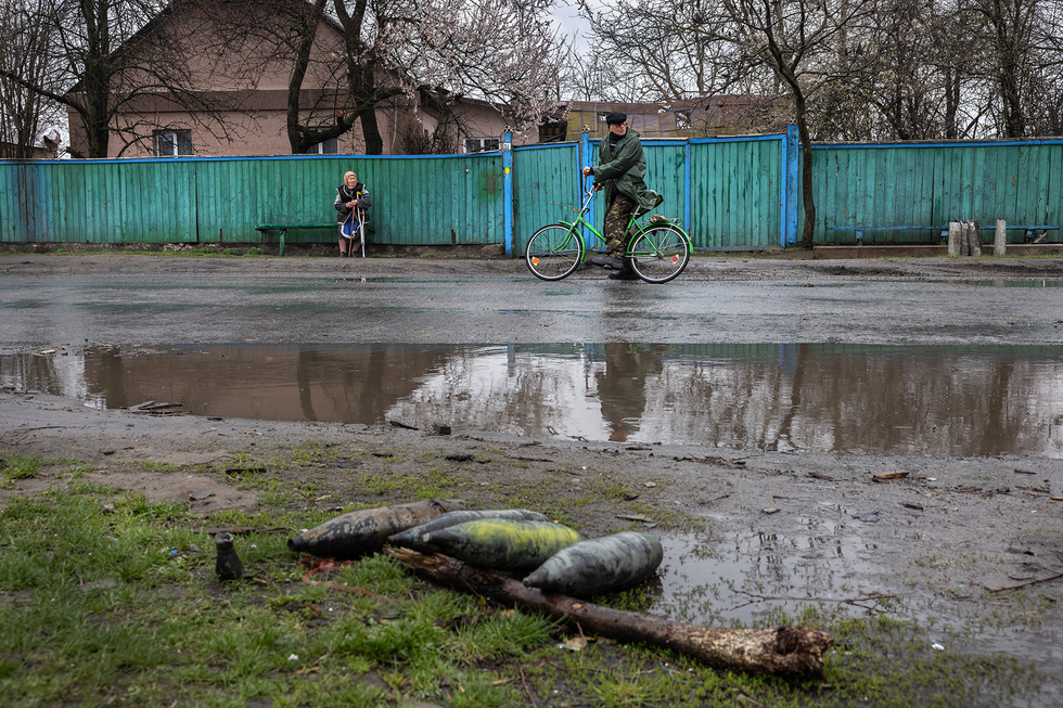 In the foreground, four pointed metal objects lie by the side of a wet and muddy road. In the background, a man rides a bicycle and a woman sits on a bench