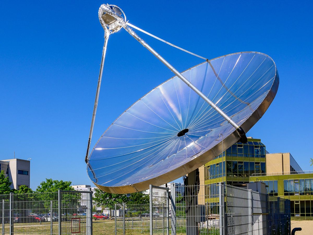 large metal segmented satellite dish with blue sky and buildings, grass and a fence behind
