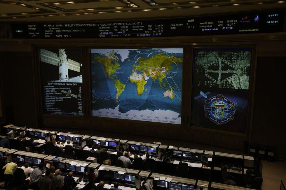 Large screens show a blue and green world map and close ups of space vehicles in front of rows of people in front of computer screens.
