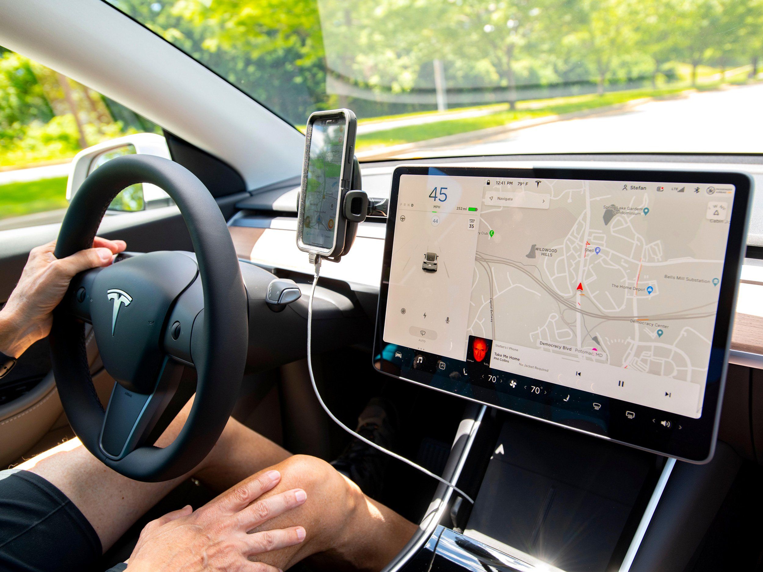 One hand sits on the steering wheel inside of a Tesla electric car. In the foreground, a map and navigation is seen on a display.