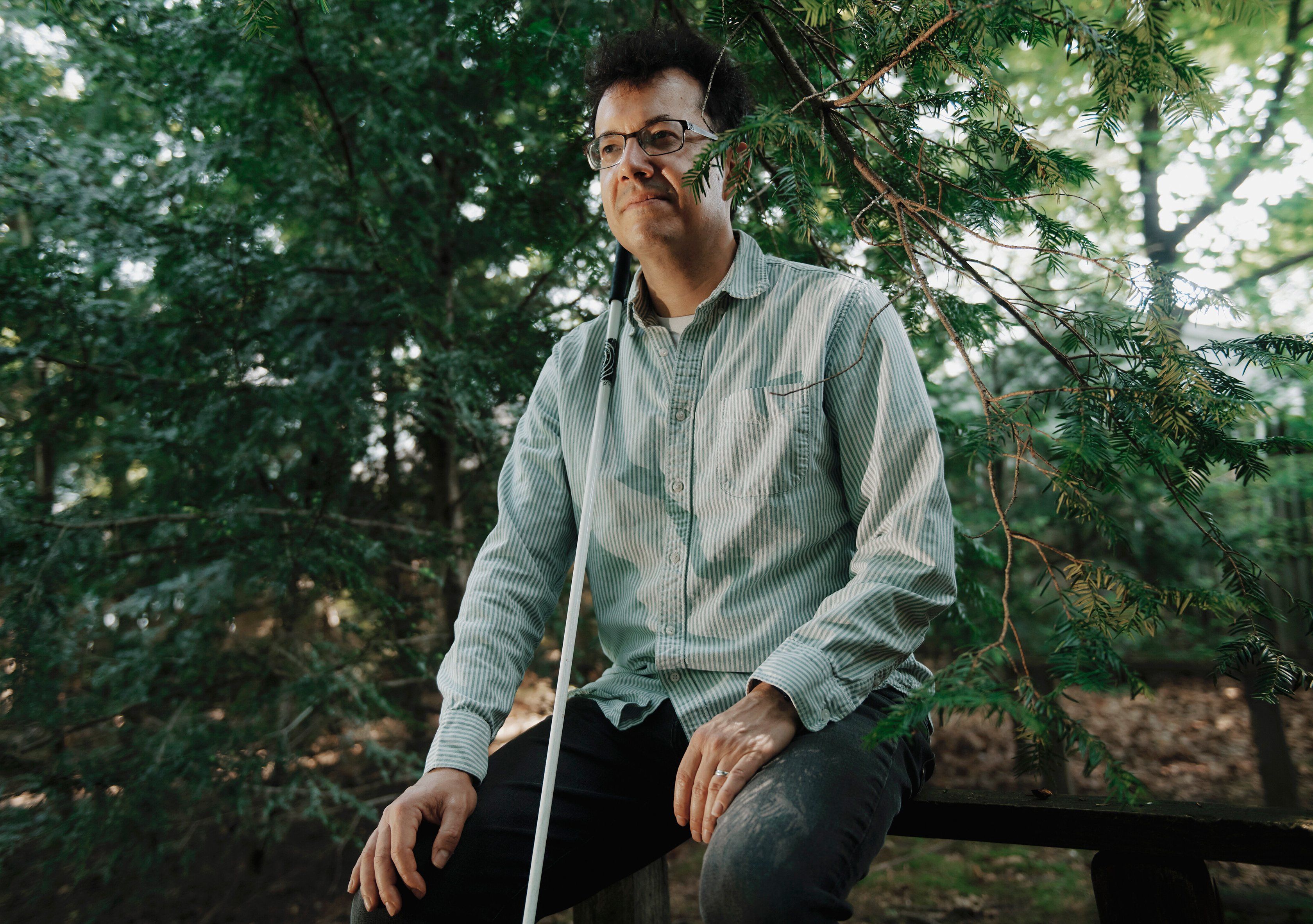 A dark haired man in glasses looks off into the distance. He sits on an outdoor bench with lush green trees behind him. A white cane rests vertically against his chest.
