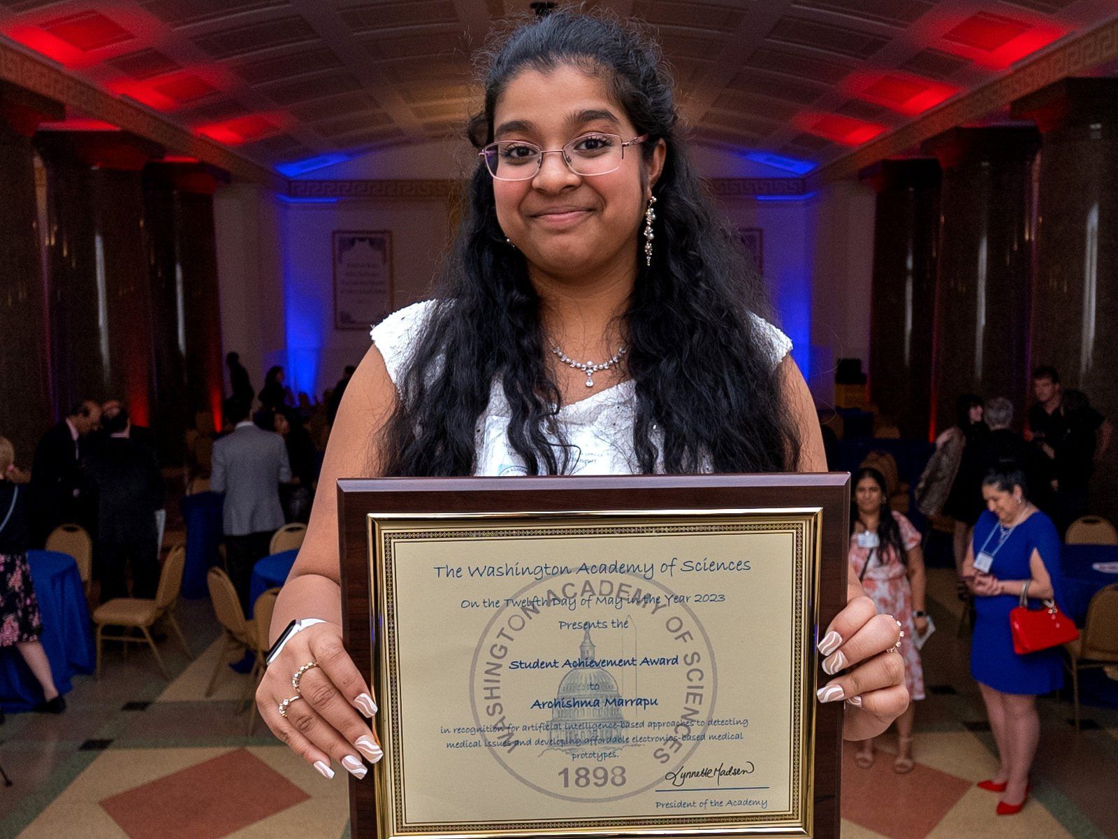 young woman smiling at camera holding a framed document with a large room filled with people in the background