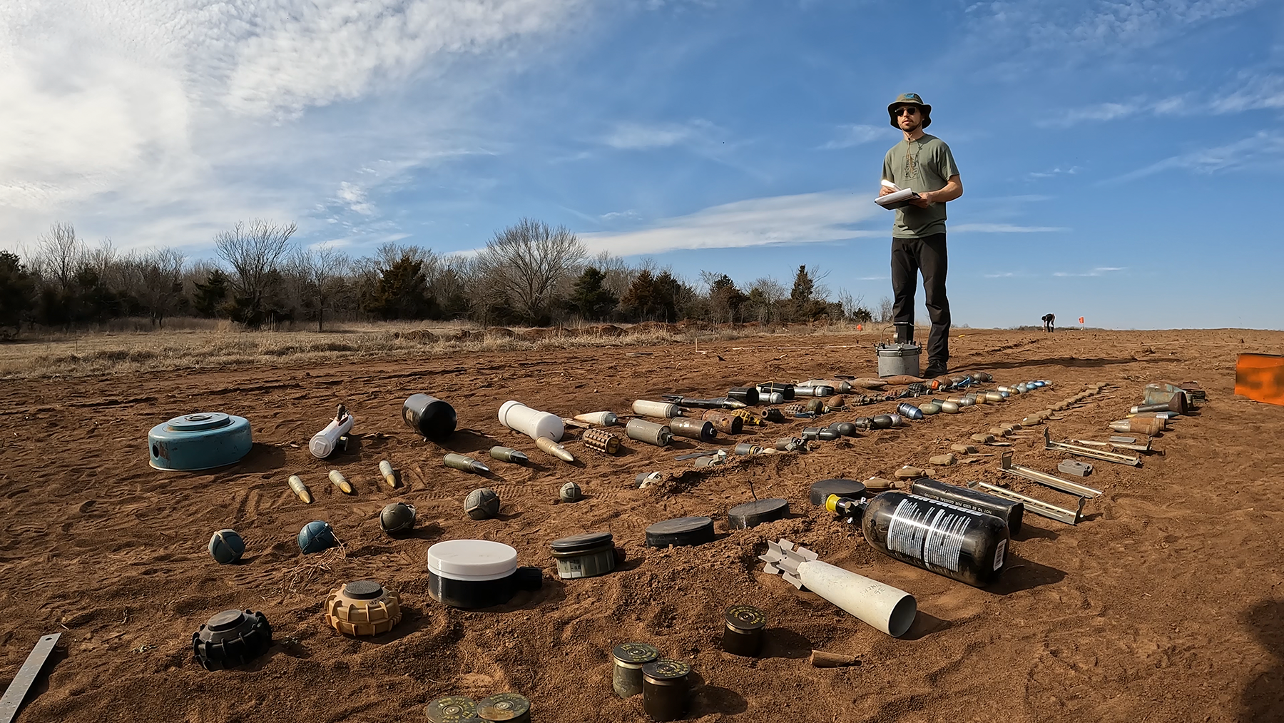 A man stands in a field with several dozen objects of different shapes and sizes lying on the ground in front of him.