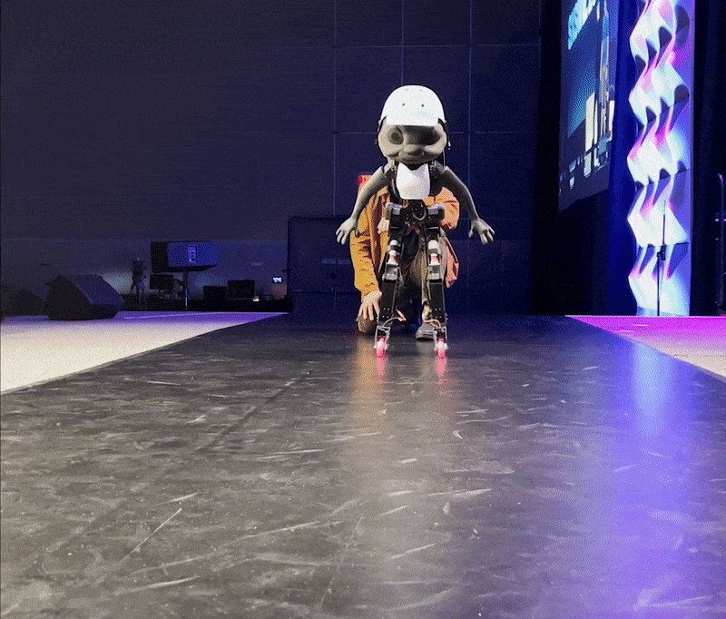 An animated gif of a grey humanoid robot in a helmet, knee pads and pink skates falling, doing a roll, and getting back up.