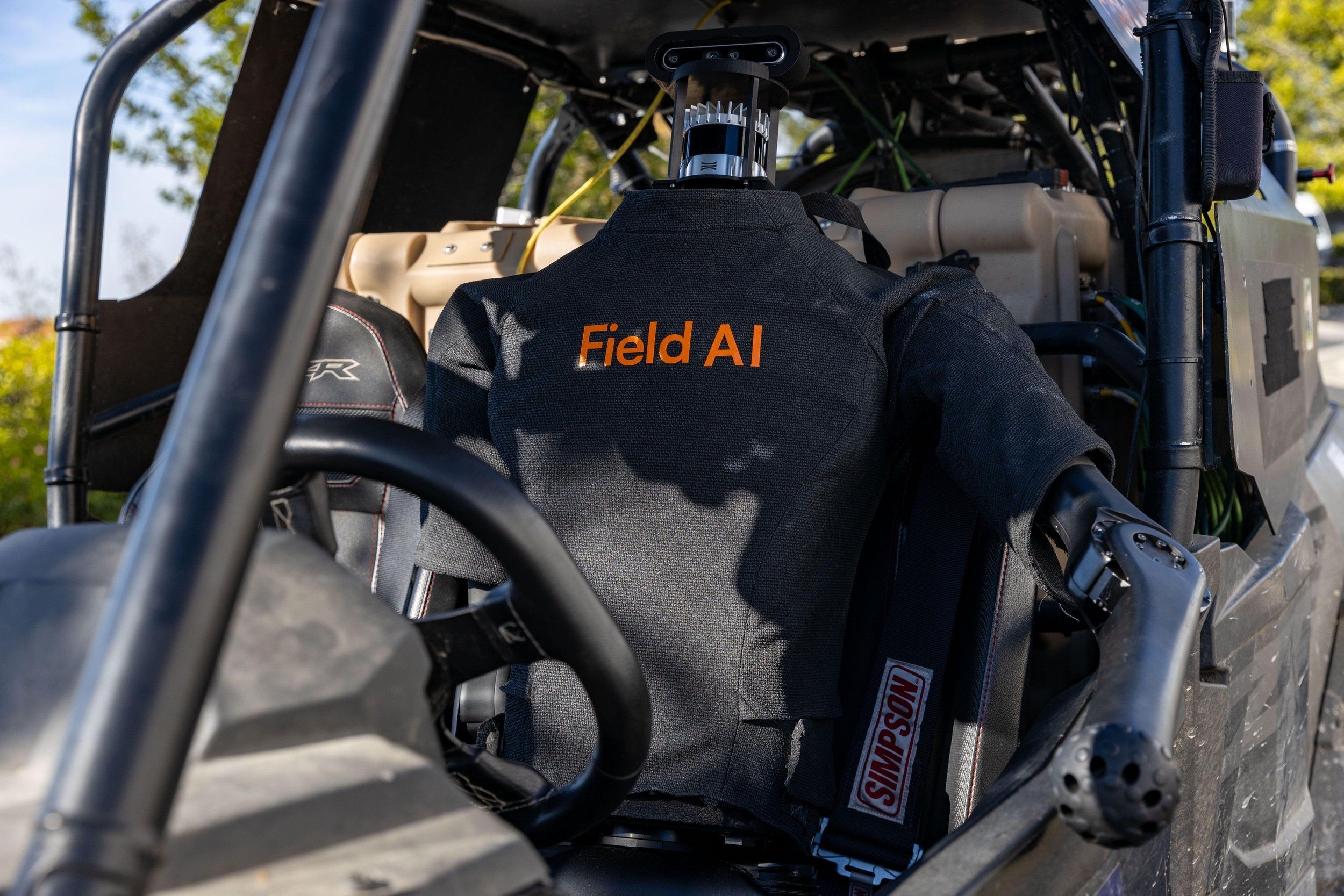 Human-like robot driver with a logo on its chest saying "Field AI" sits behind the wheel of an open, dune buggy-like vehicle