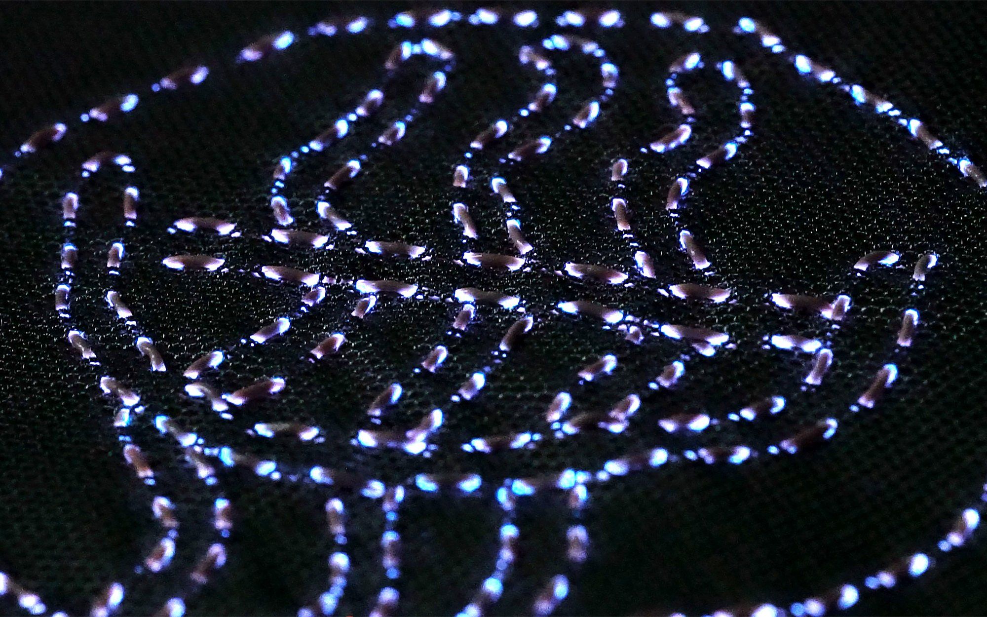 Glowing tubes emit a purplish light, and are woven into a black fabric in a pattern of wavy lines and a circle
