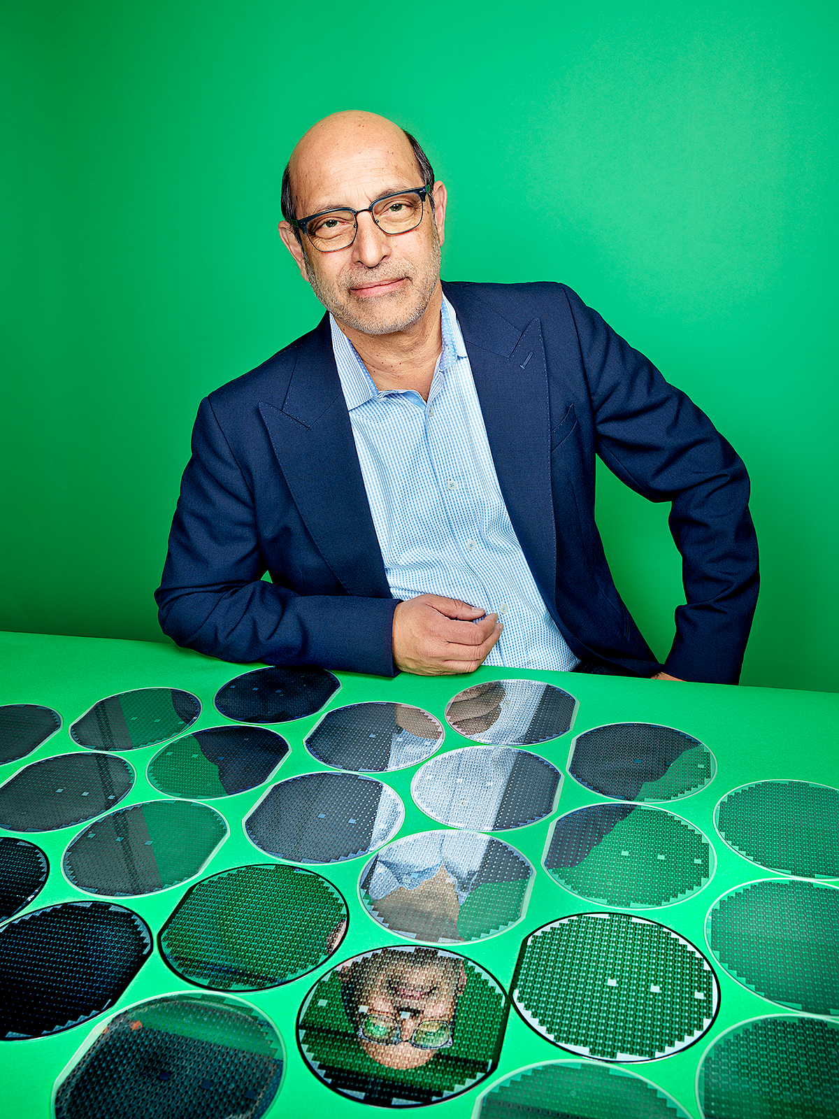 Photo of a man in glasses and jacket sitting in front of semiconductor wafers.
