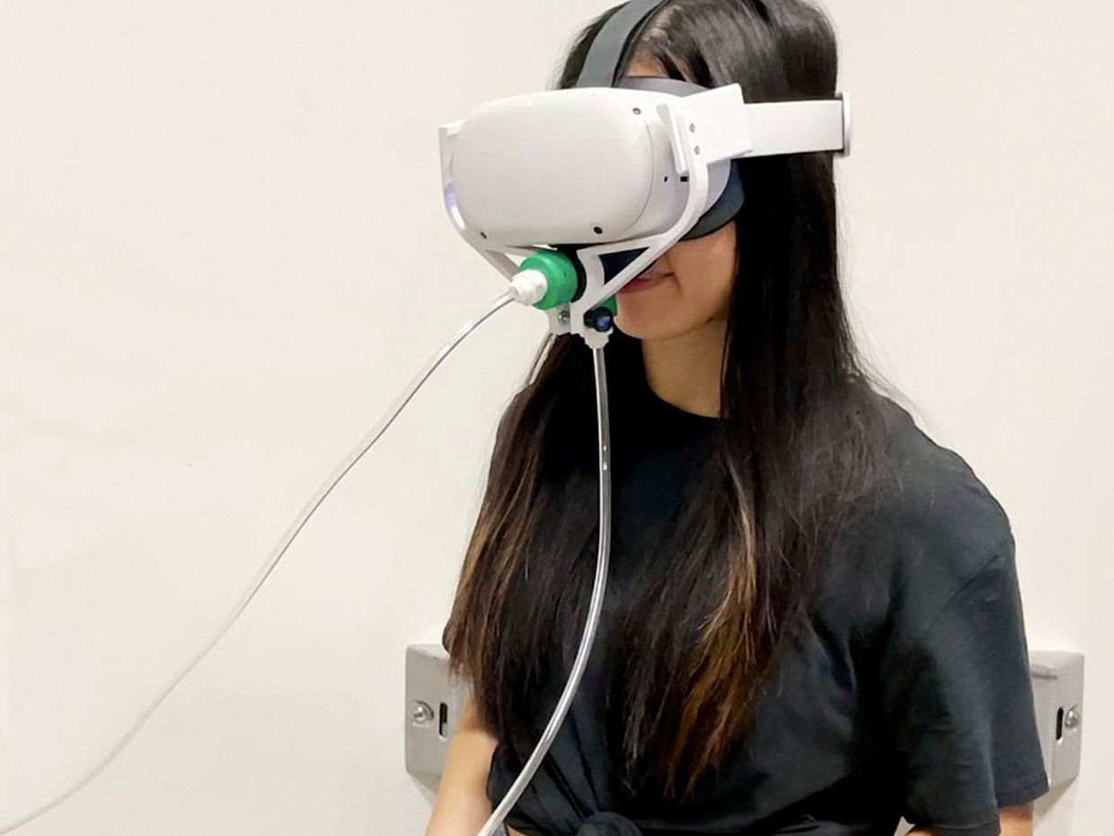 A person with long hair wearing white VR googles with a tube connected to the nose area against a white background