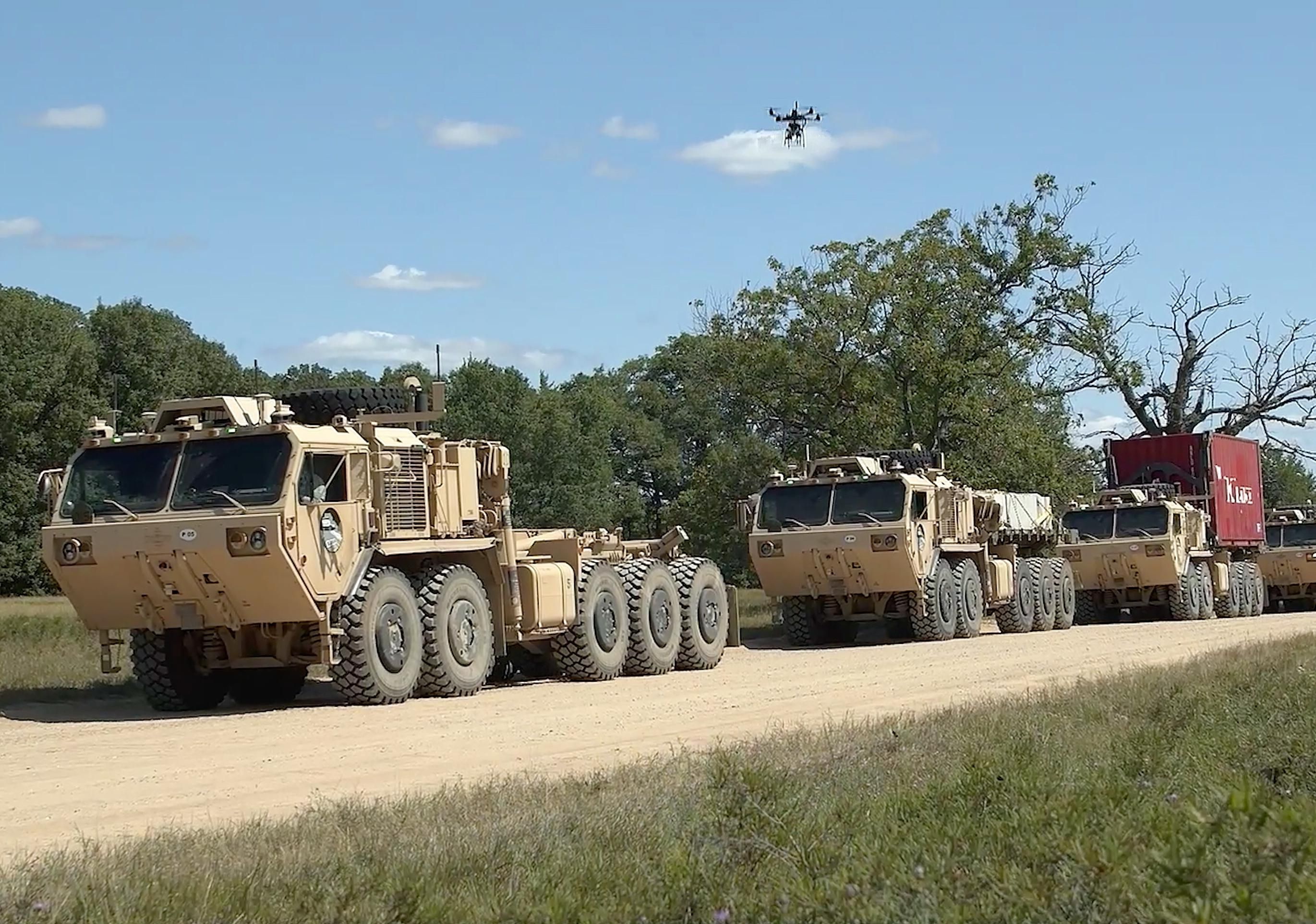 4 large military vehicles on a dirt road. The third carries a red container box. Hovering above them in a blue sky is a large drone.