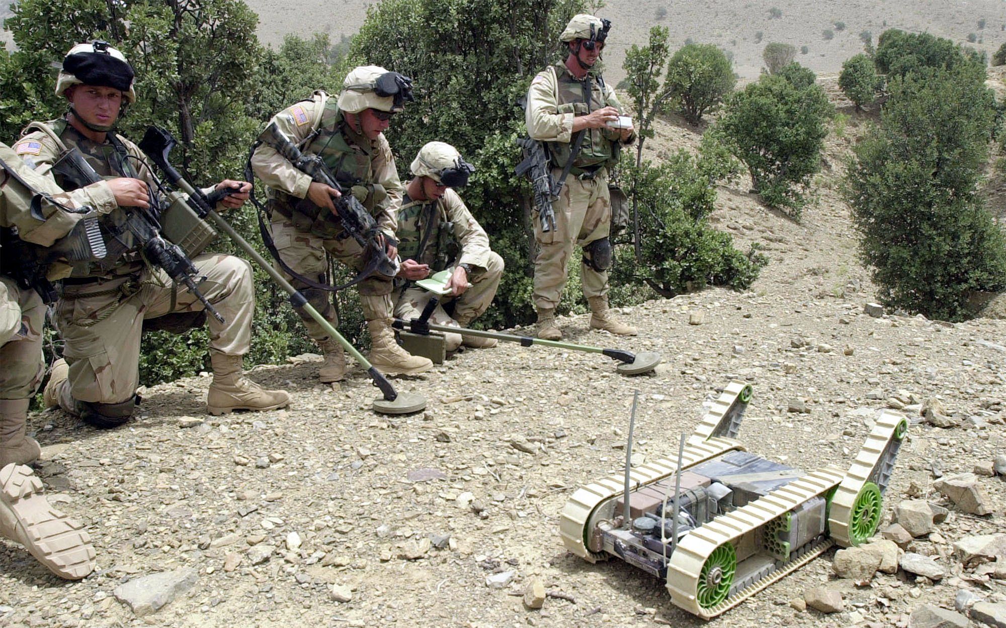 Four men wearing army fatigues and carrying weapons and tools look on as a small robot with treads rolls along the dusty ground.