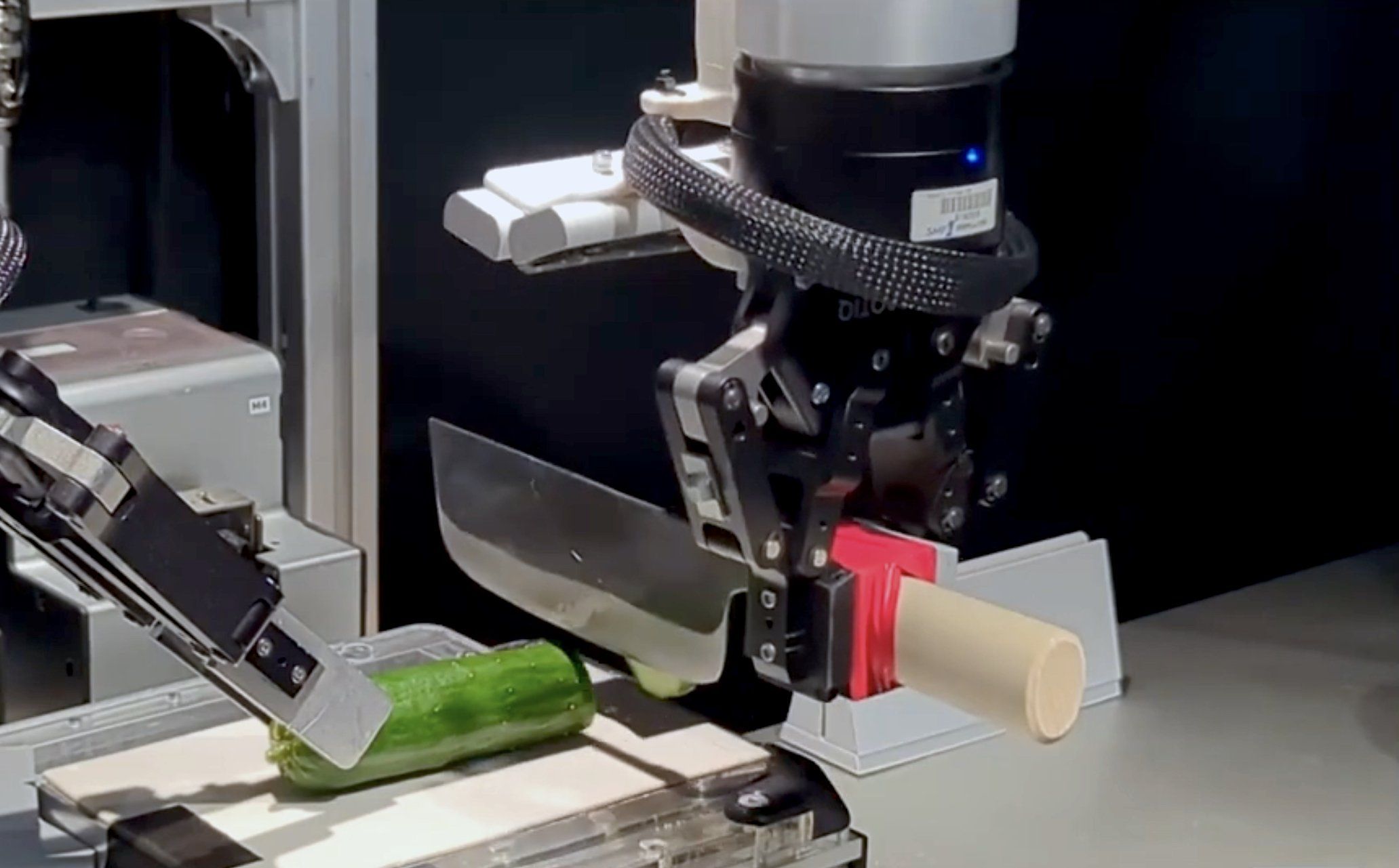 A robotic hand uses a kitchen knife to slice a cucumber.
