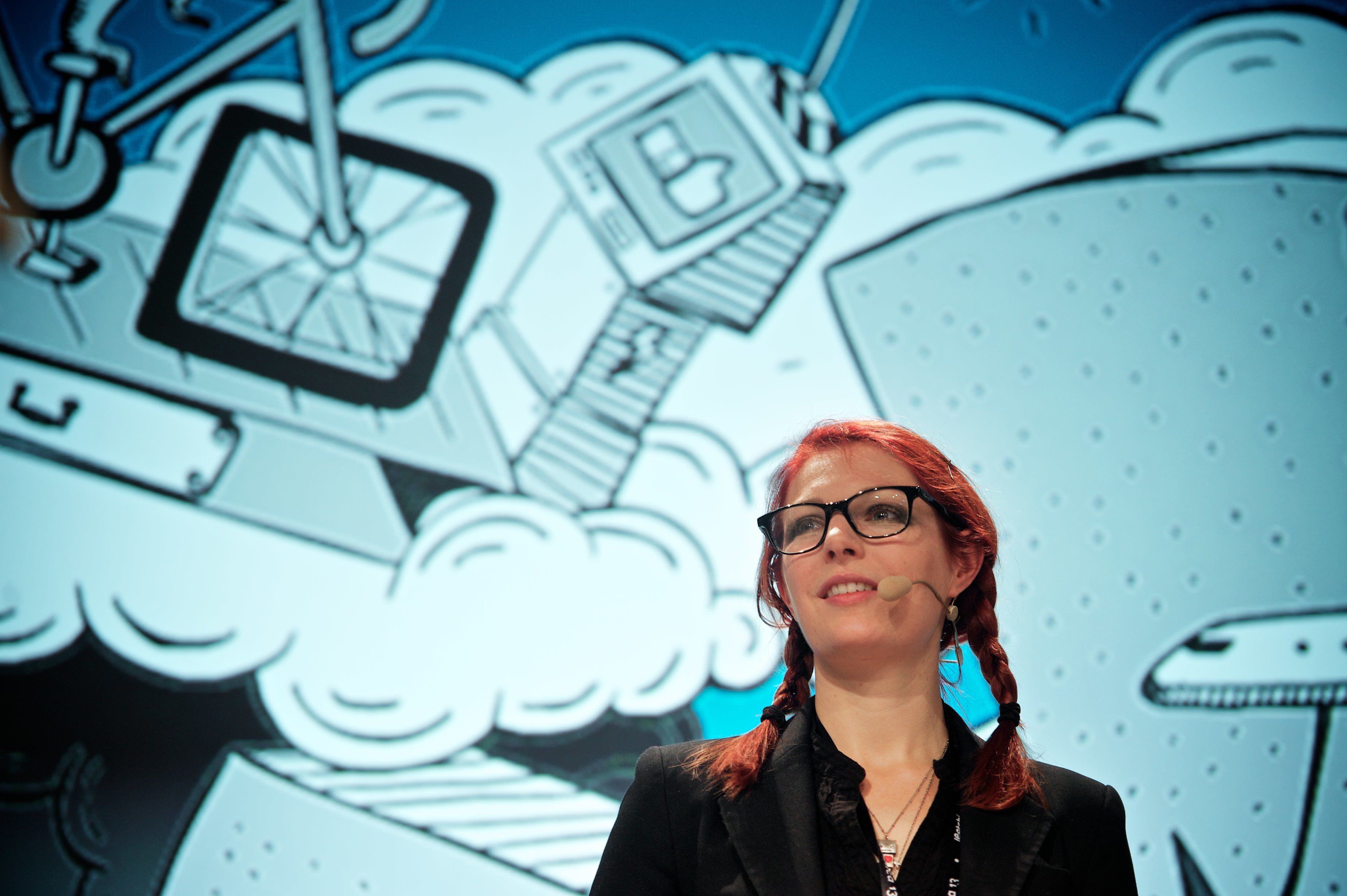 A woman with red colored hair, dark rimmed glasses, and black jacket, speaks at a conference in front of a screen showing comic book style illustrations of robotic contraptions.