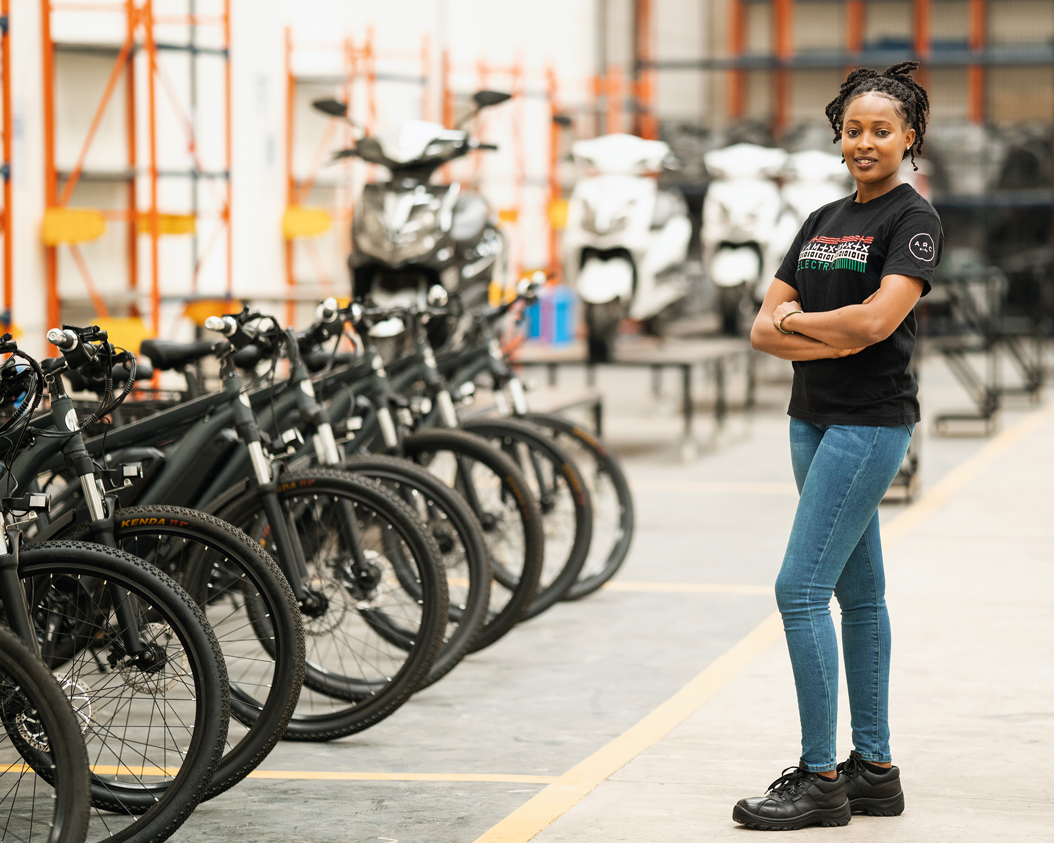 A woman dressed in a black T-shirt and blue jeans standing inside a building in front of a row of electric bicycles.