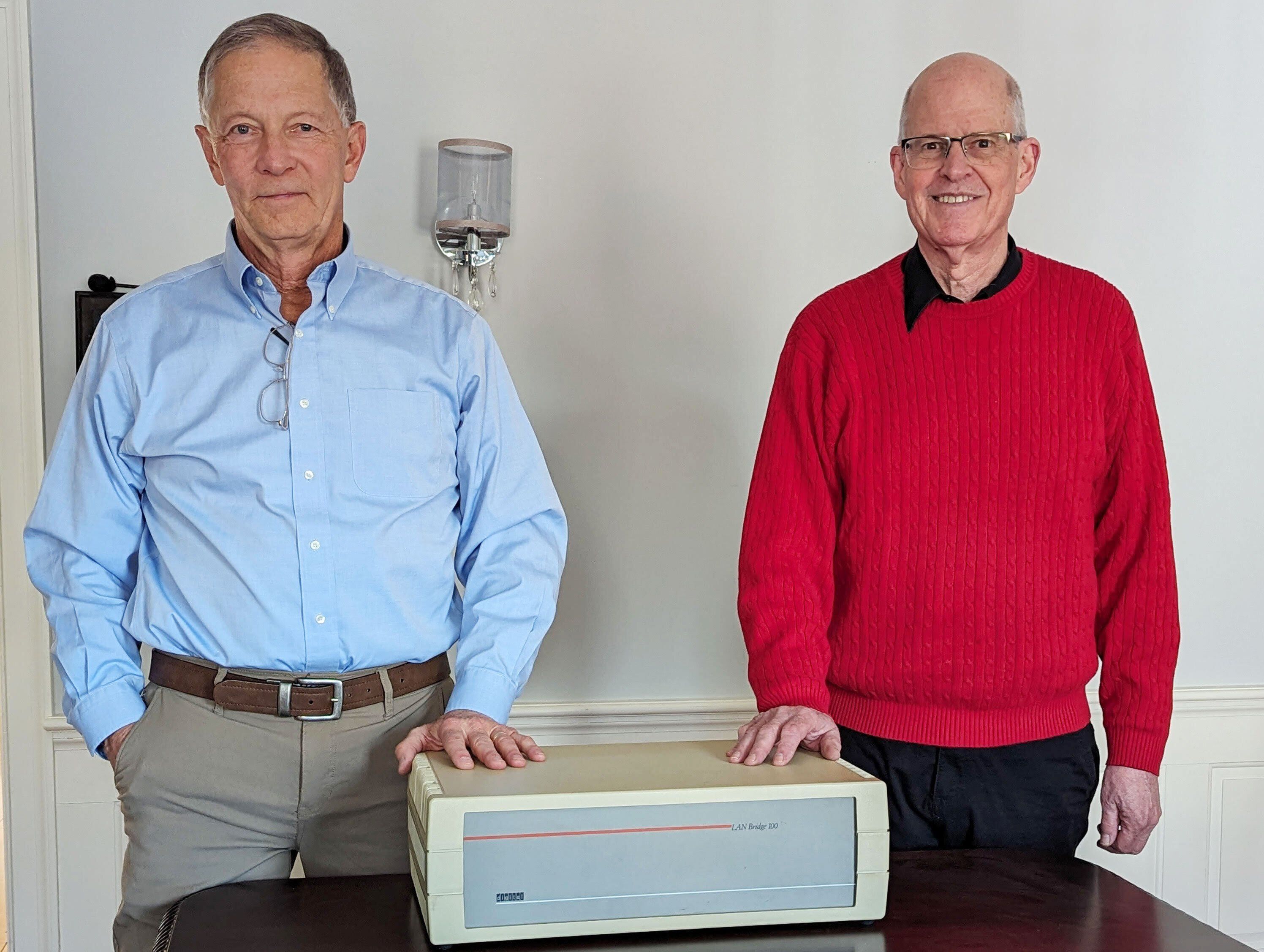 portrait of two men standing with their hands on a small beige and gray box while looking at the camera