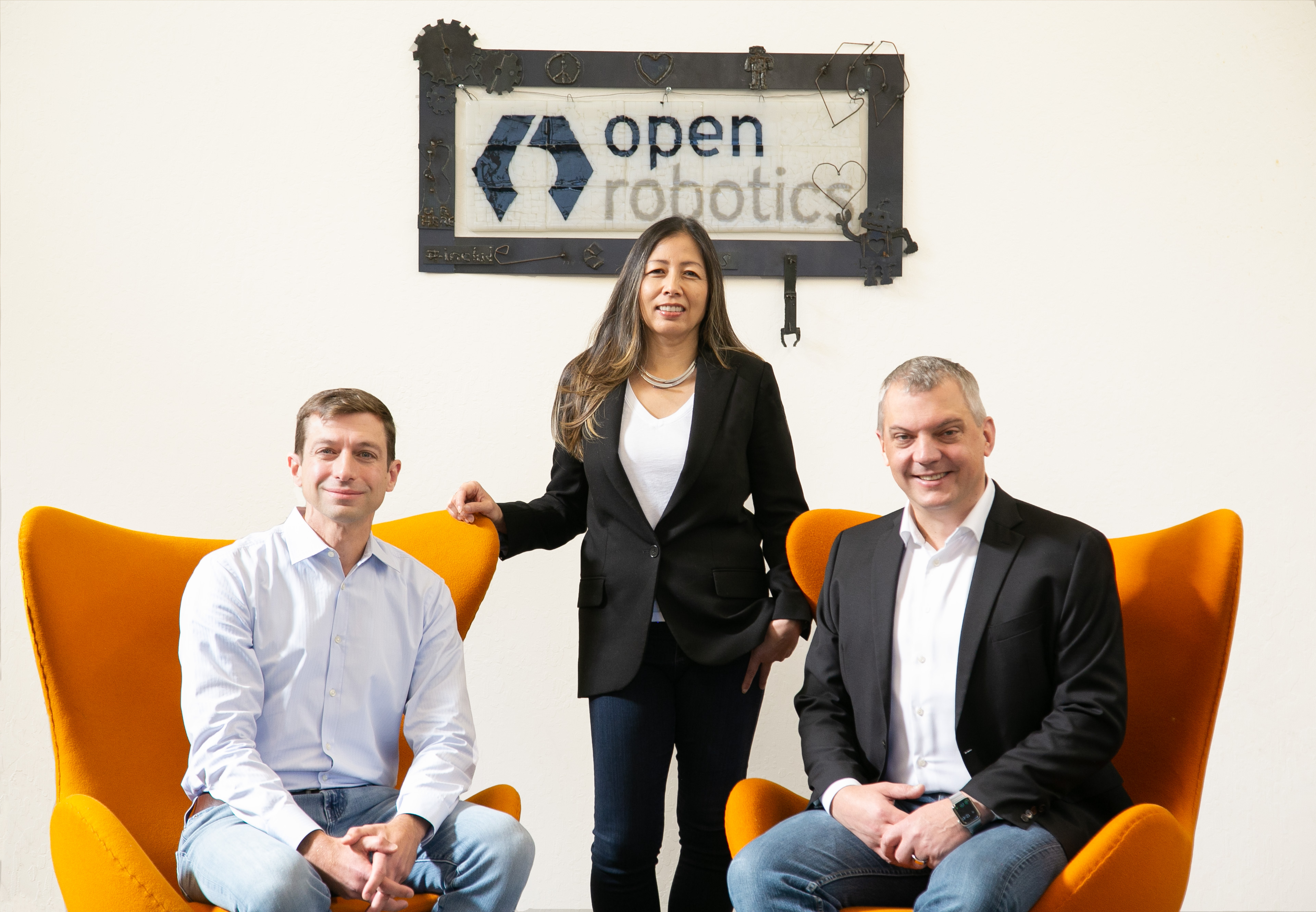 A woman stands behind two men sitting in orange chairs under a sign that says "Open Robotics"