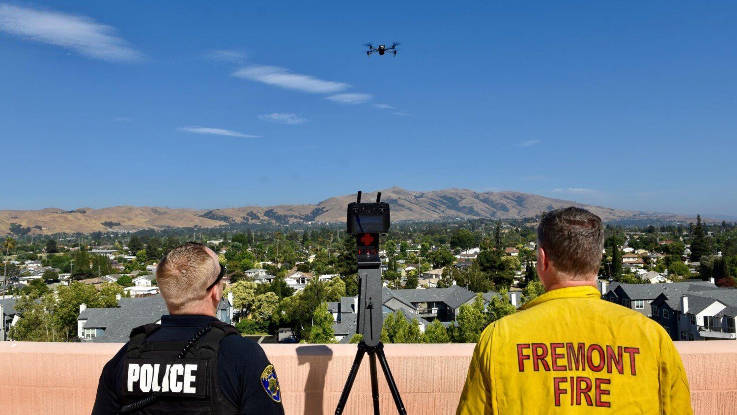 a police officer and a fire fighter standing on top of a building with their backs facing the camera watching a drone fly in the sky over a city setting