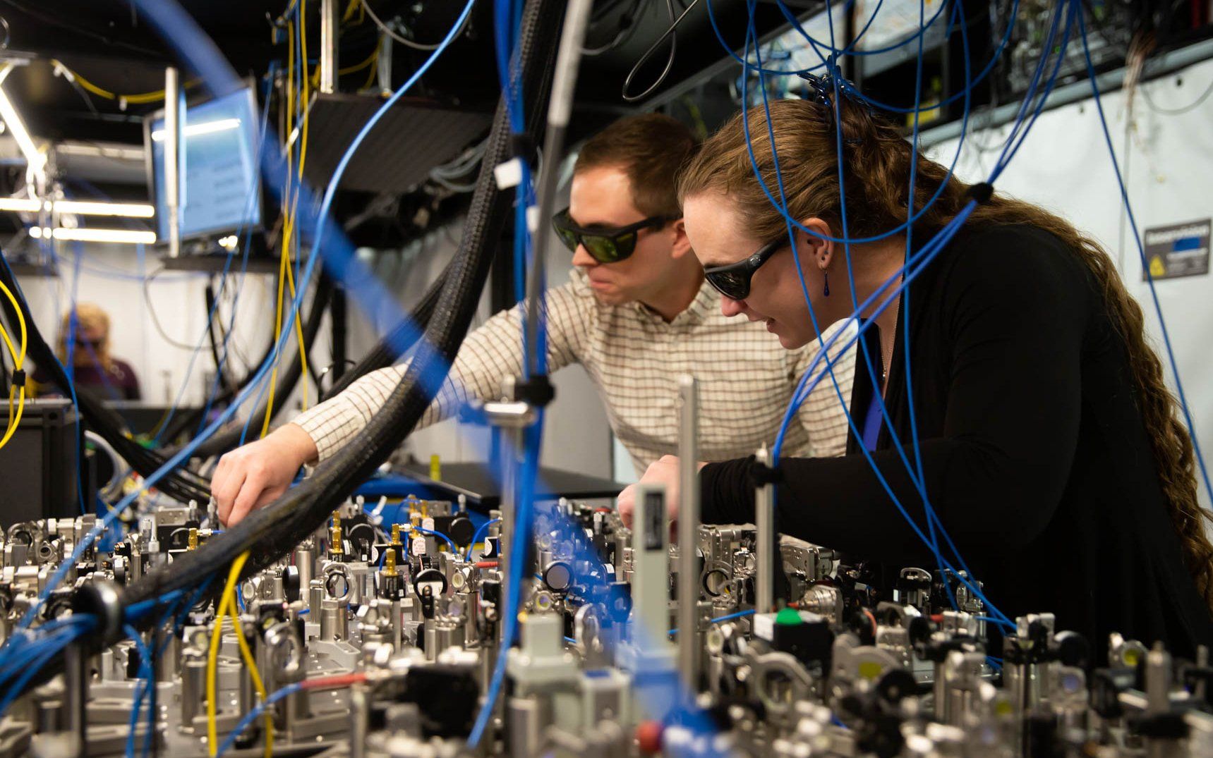 Two people in dark lab glasses stand over a complicated looking tabletop laser 