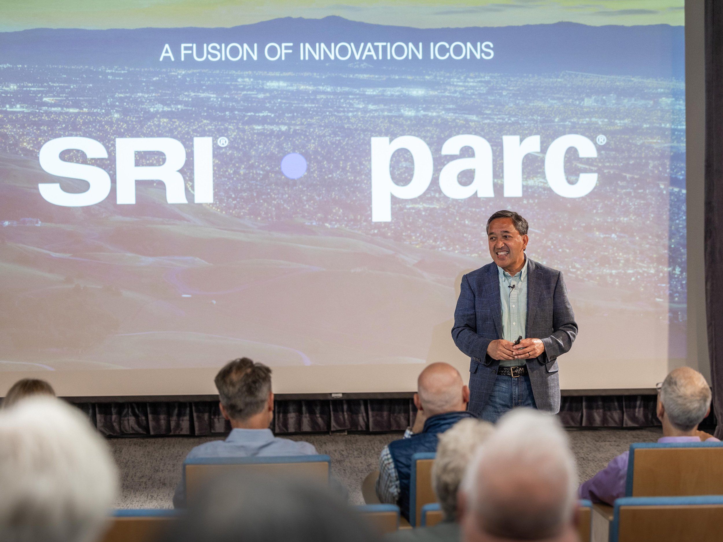 man speaking to a group of people in front of a screen that says "SRI parc" and in smaller letters "A fusion of innovation icons"