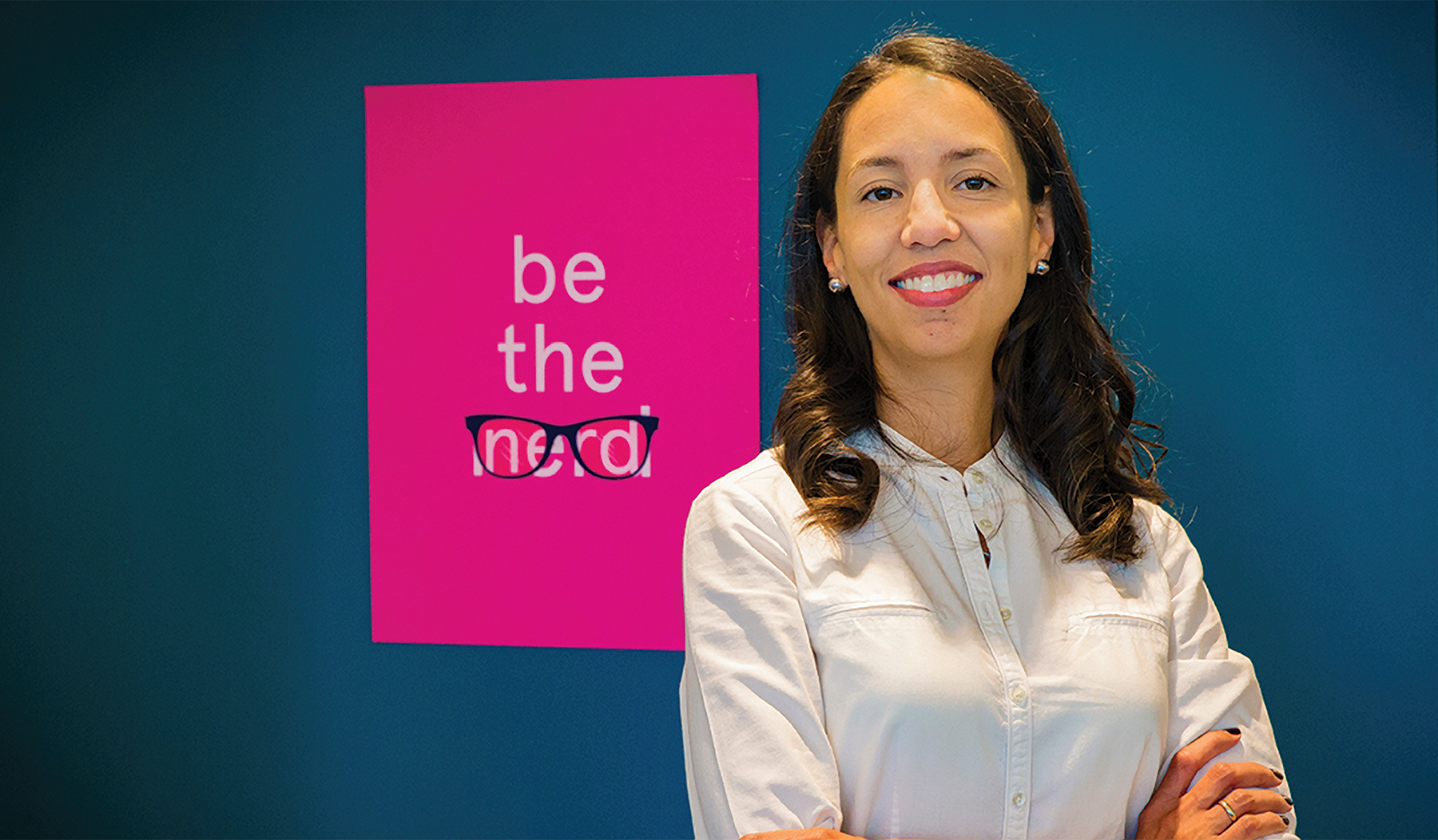 A photo of a smiling woman in front of a pink sign that says, "be the nerd."