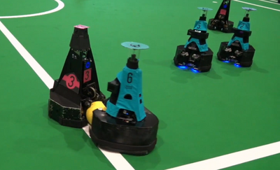 Five waist-high pyramidal mobile robots squabble over a yellow soccer ball on a green soccer field.