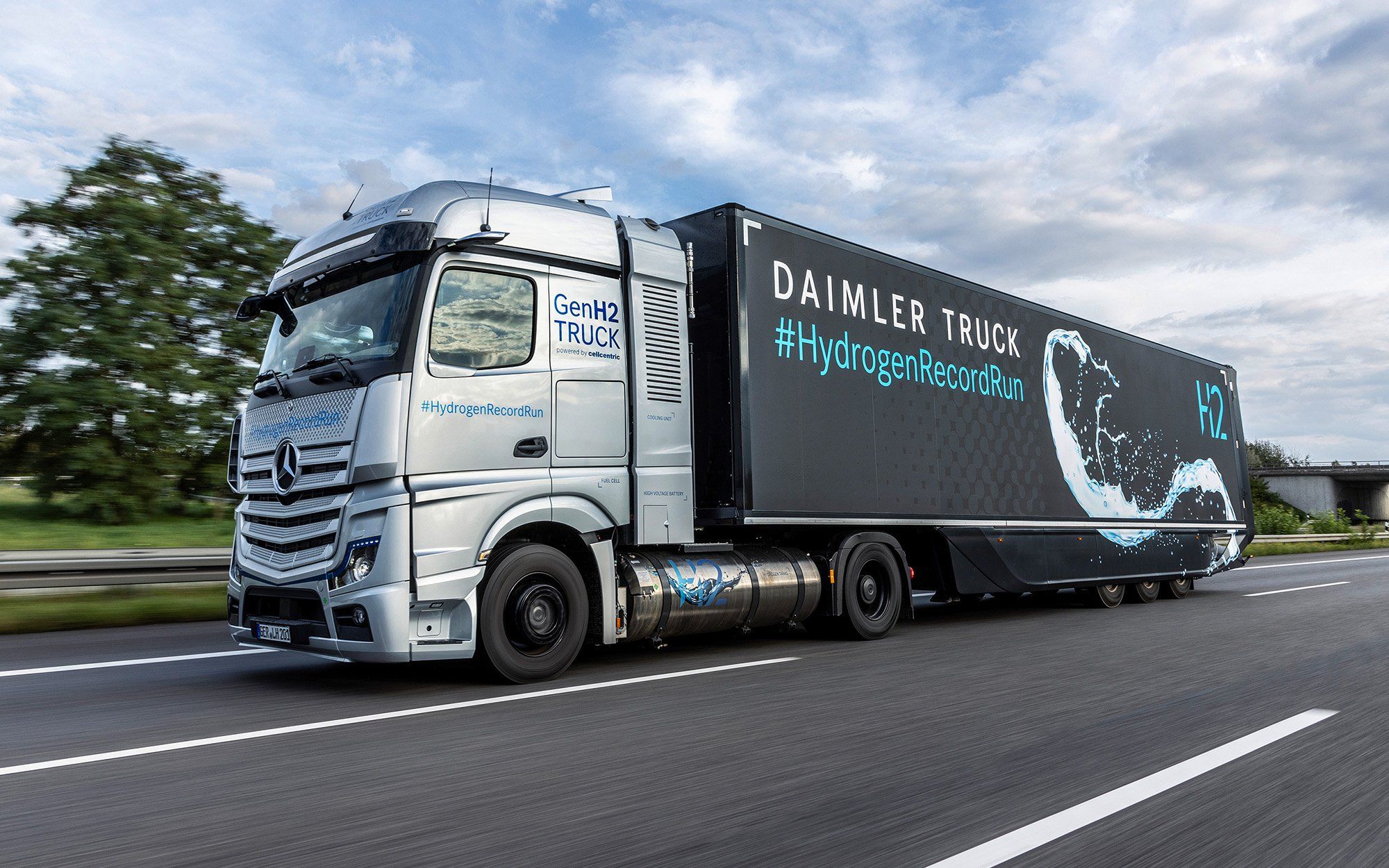 A grey and black semi truck on a highway is covered with branding for GenH2 Daimler Truck