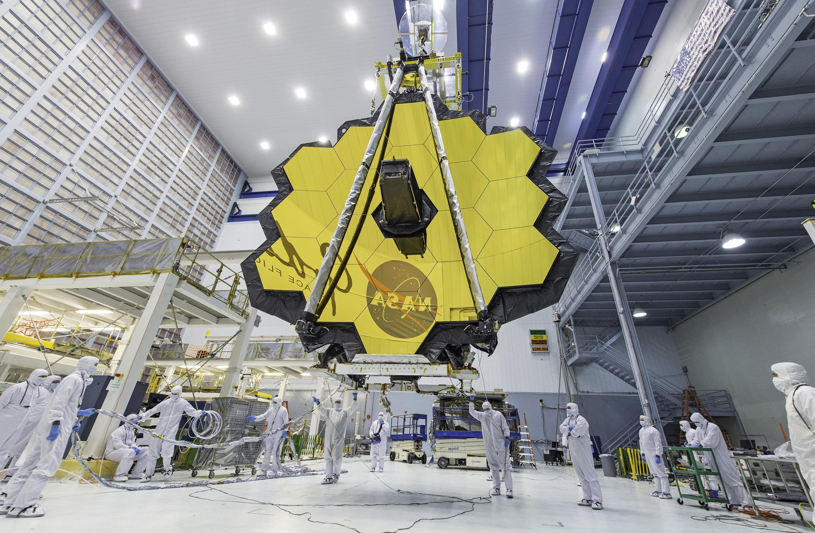 Fourteen technicians in clean-room suits guide the hoisting of a honeycombed, hexagon-mirrored telescope inside a giant cleanroom construction space 