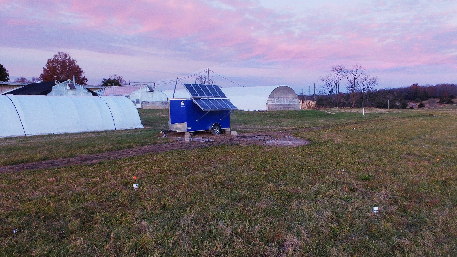 A grassy field with scattered white boxes in the ground, a blue trailer with solar panels, and multiple white greenhouse tents. 