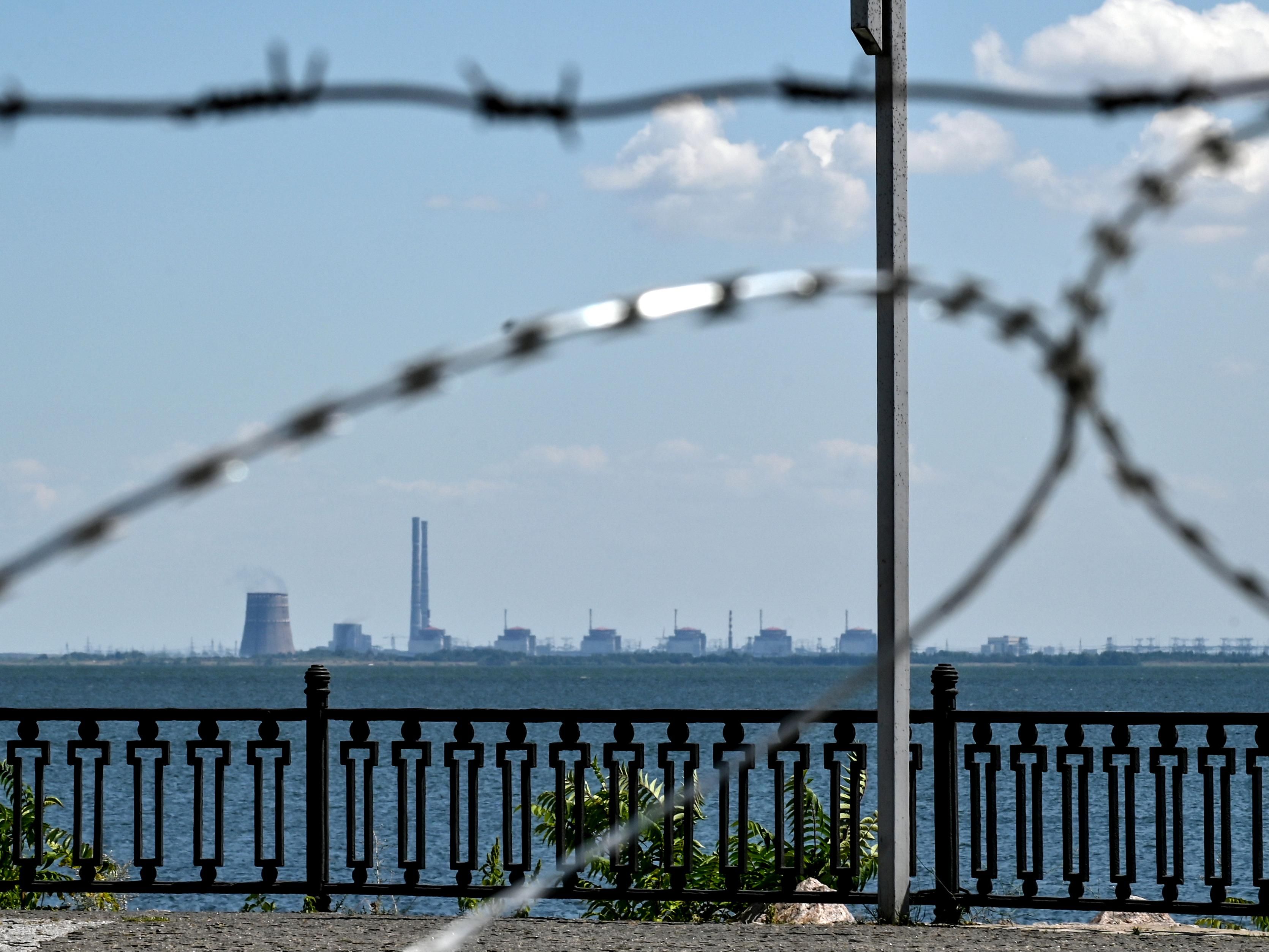 Barbed wire in the foreground frames a power plant in the distance across a wide river.