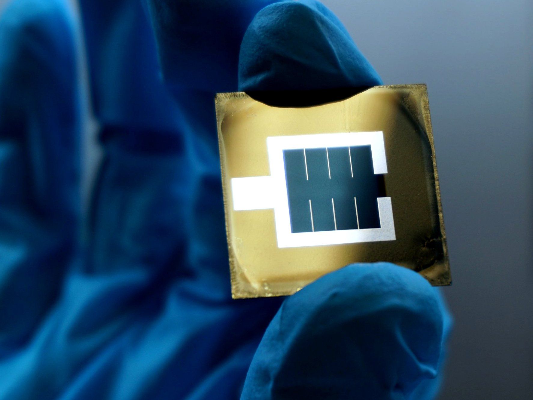Blue gloved hands hold a golden square of material with a blue rectangle of solar material surrounded by white lines.