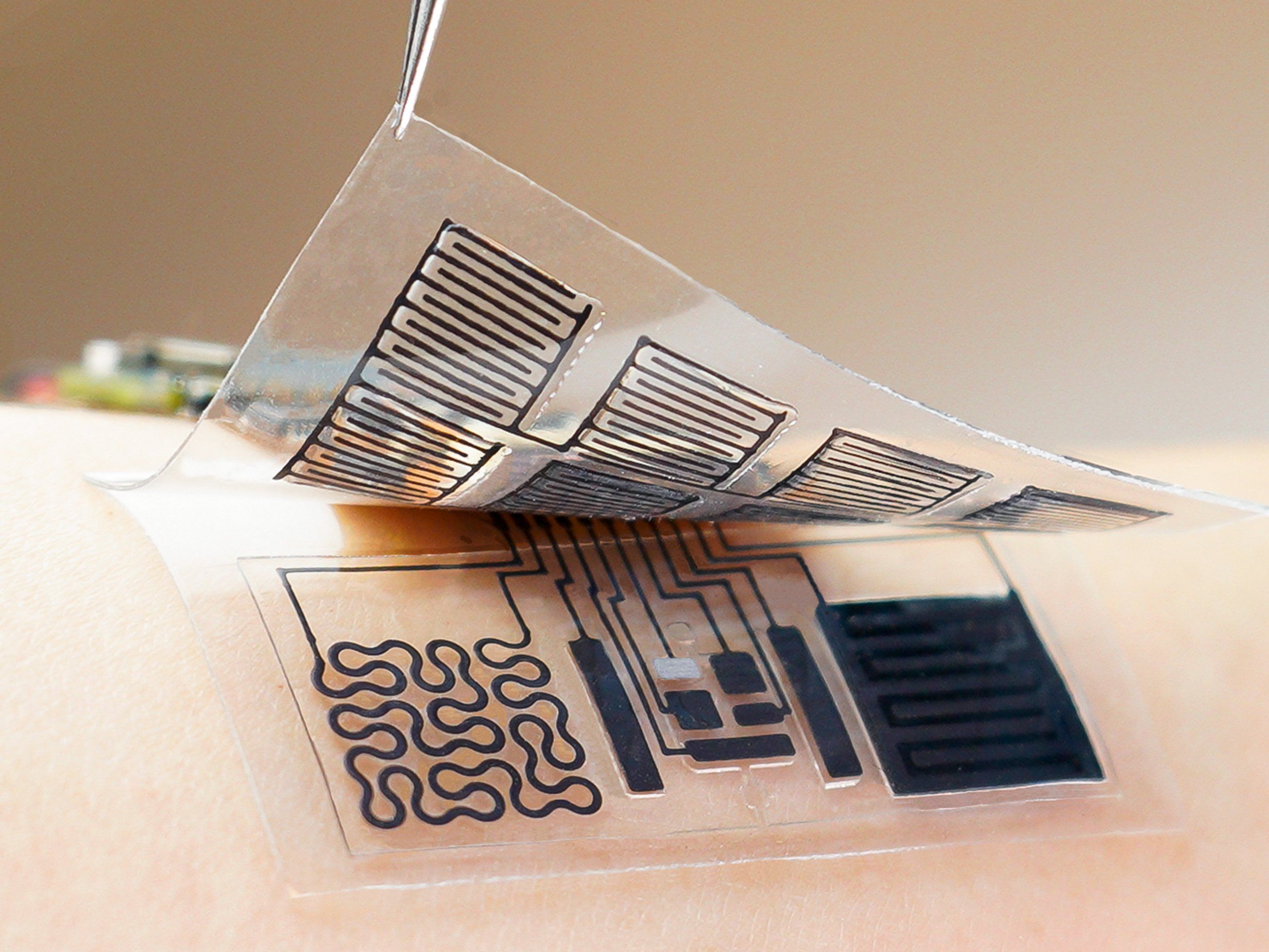 A piece of material with a black circuitry pattern is pealing off a second piece, with an intricate electronics pattern, which sits on a person's skin.