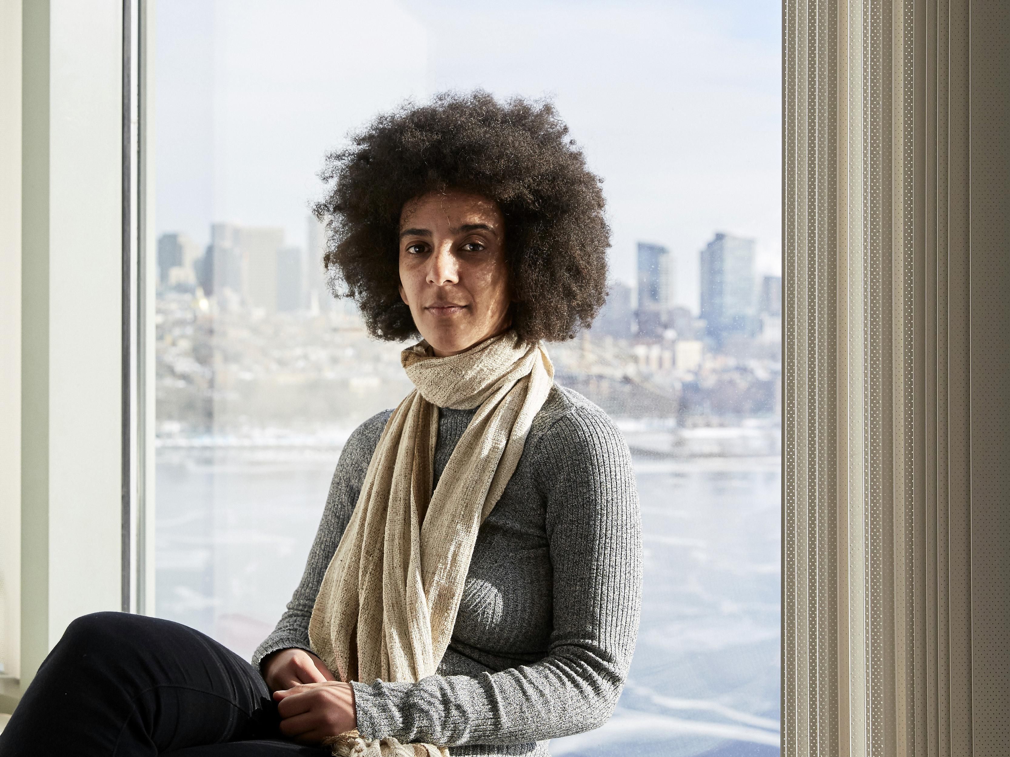 Portrait of a woman in a grey sweater and cream colored scarf sits in front of a window