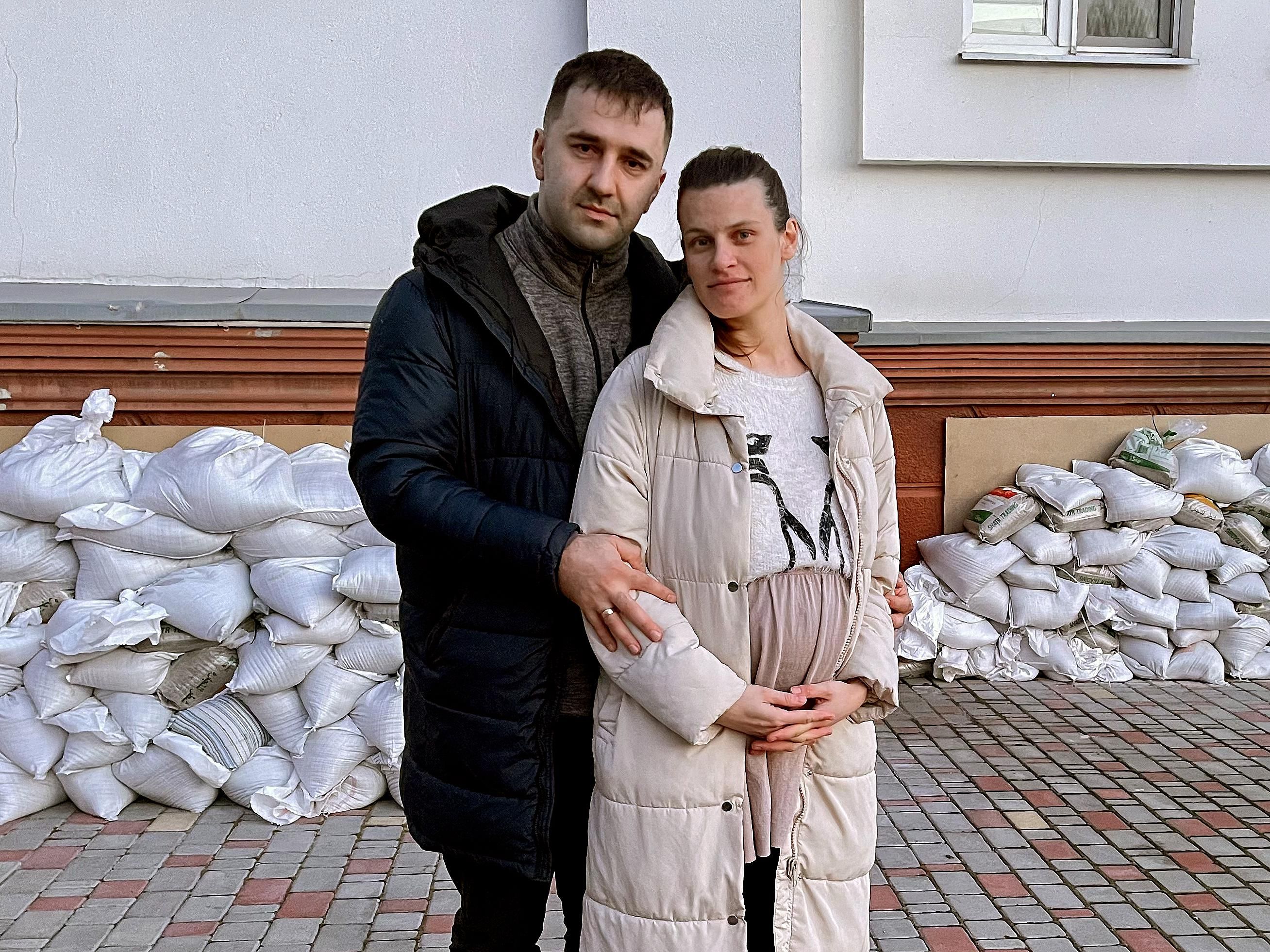 A man and a pregnant women pose outside in front of a pile of sandbags.