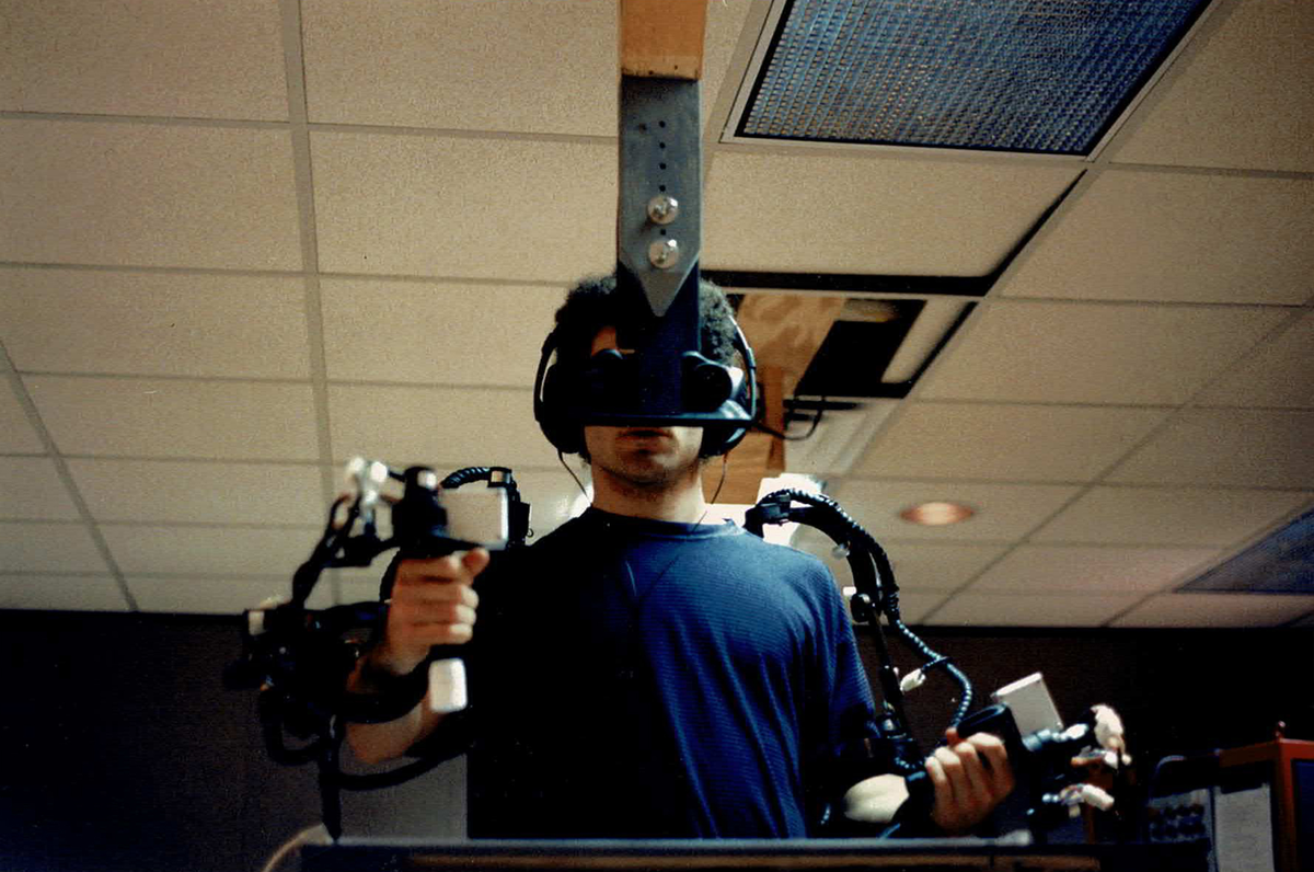 Man wearing goggles and earphones, holding wired controllers in each hand. 