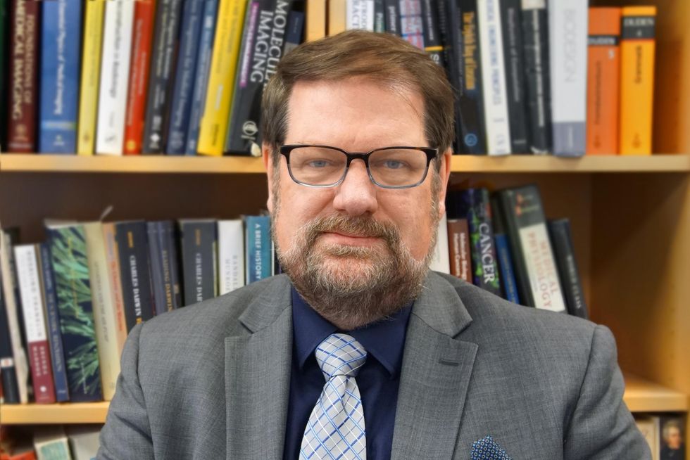Man with beard and eyeglasses in a suit against a background showing bookshelves.