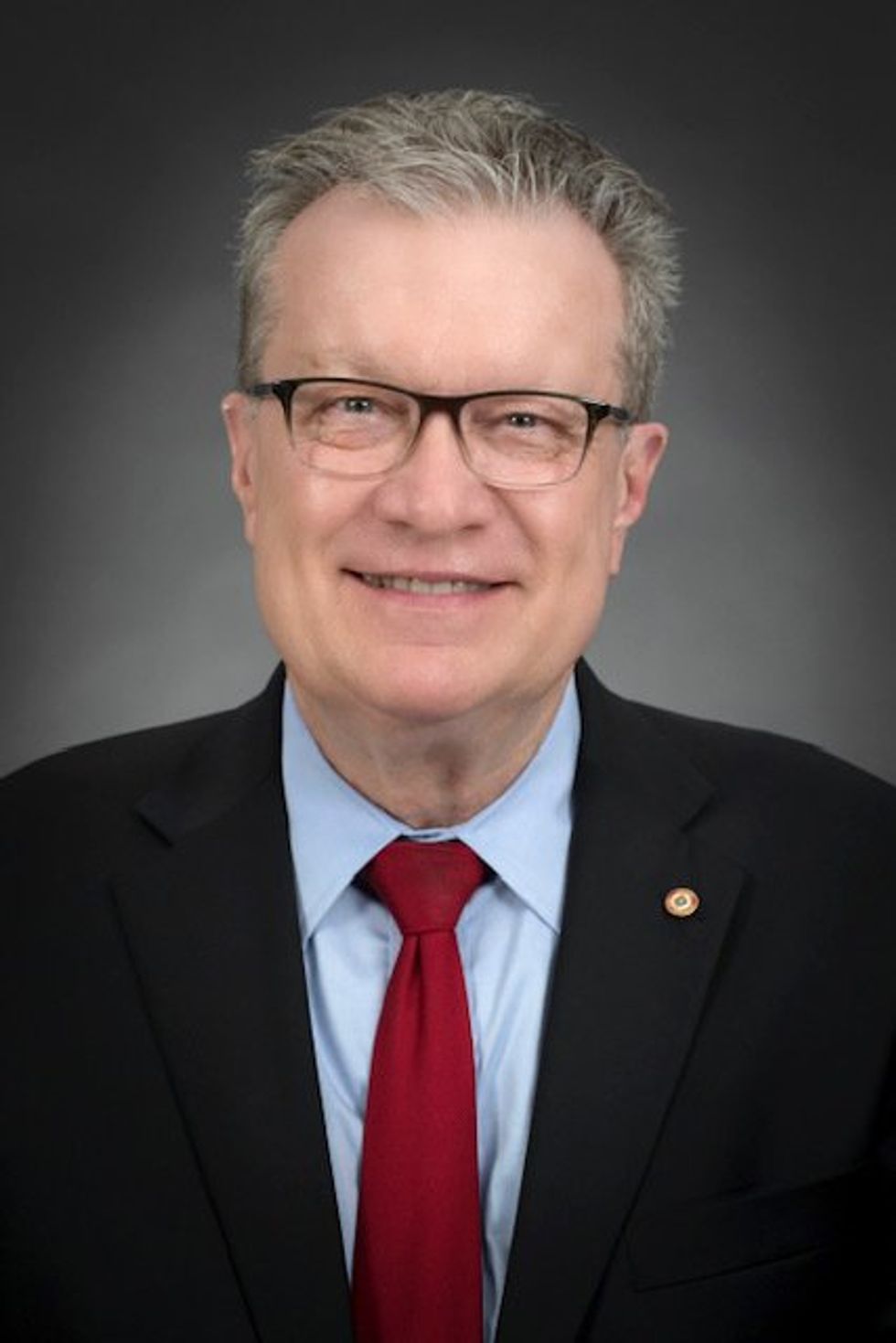 man with glasses wearing a red tie, blue shirt and black blazer in front of a gray background