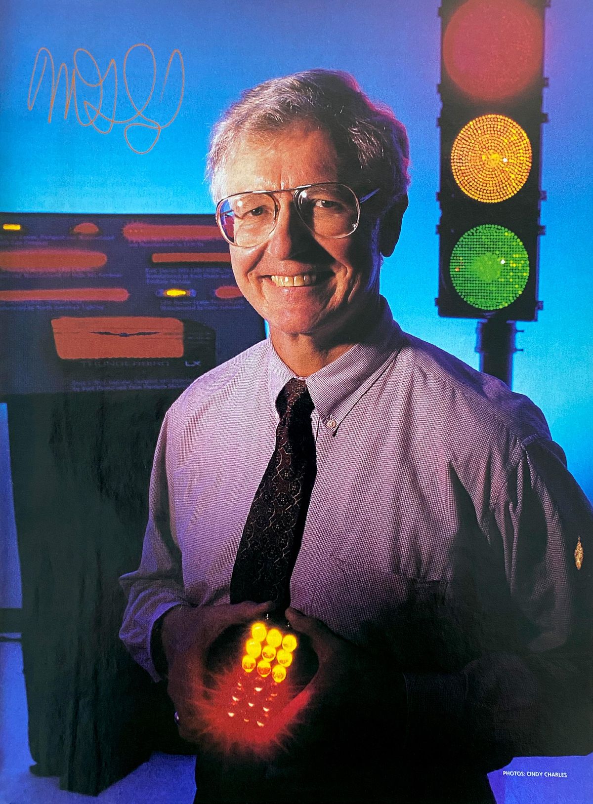 Man  with grey hair wearing dress shirt and tie standing in front of an LED stoplight and holding a panel with yellow and red LEDs glowing