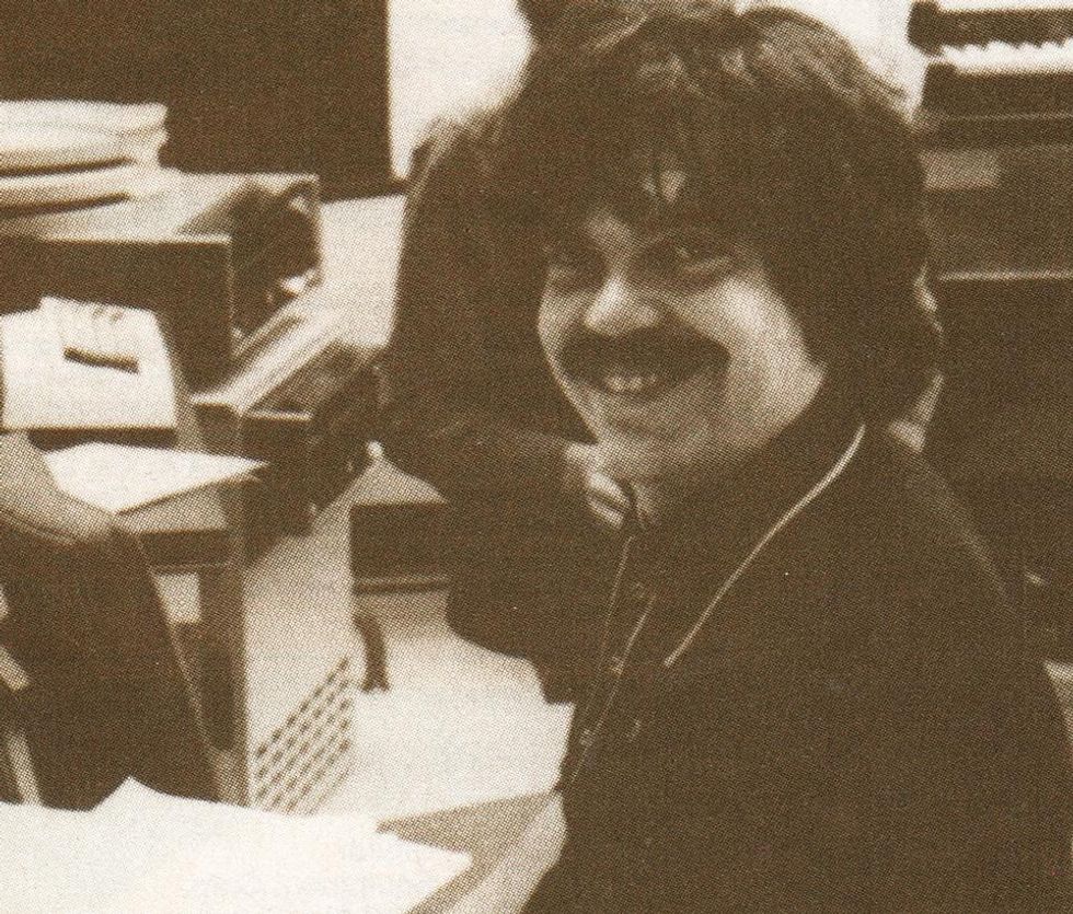 Man with long hair and mustache sitting at a desk, computing equipment behind him