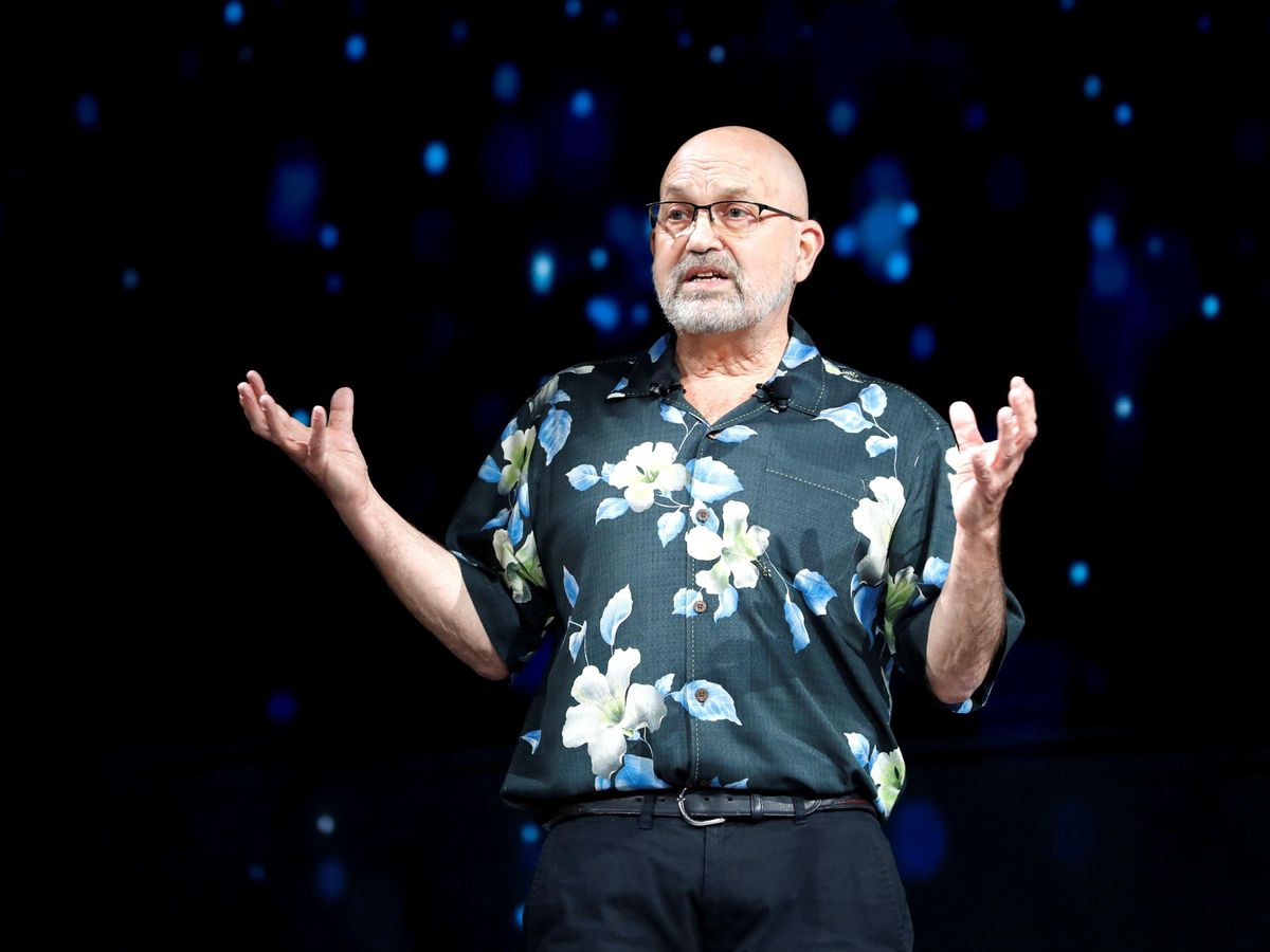 Marc Raibert, an older white man with a bald head and a short white beard and glasses, gestures as he speaks on a stage. He is wearing formal pants and a flower-print short sleeve shirt.