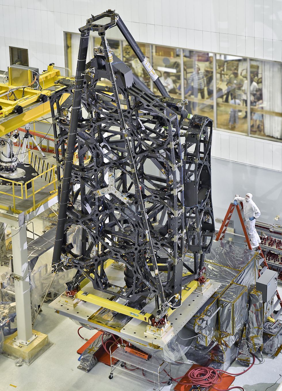 Metal superstructure of cages and supports stands on a giant platform in a warehouse-sized clean-room. A man in a cleanroom suit watches the operations.