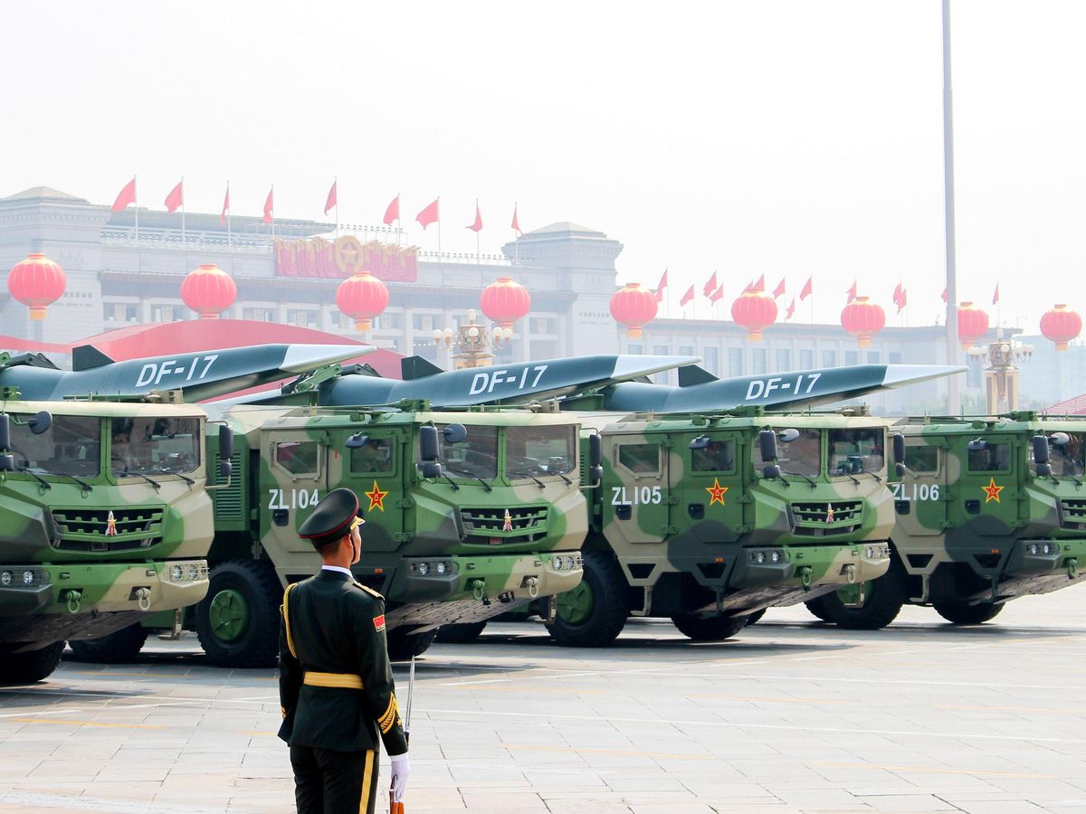 Military guard stands sentry in front of four camouflaged trucks, armed with large missiles on their roofs