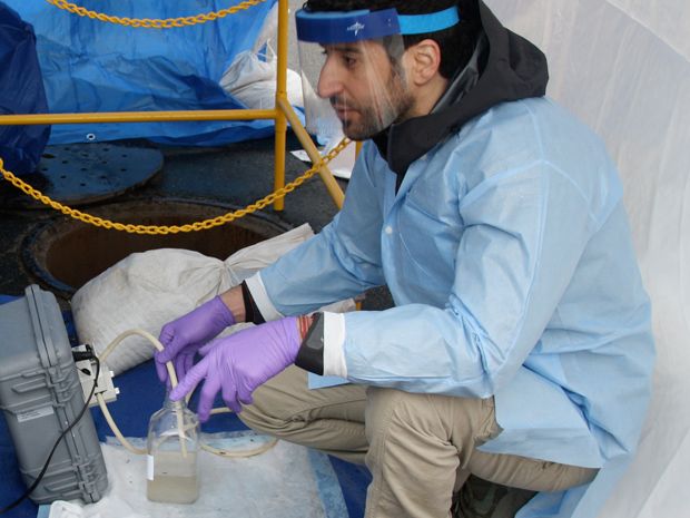 Man in blue coat and face mask sampling sewage.