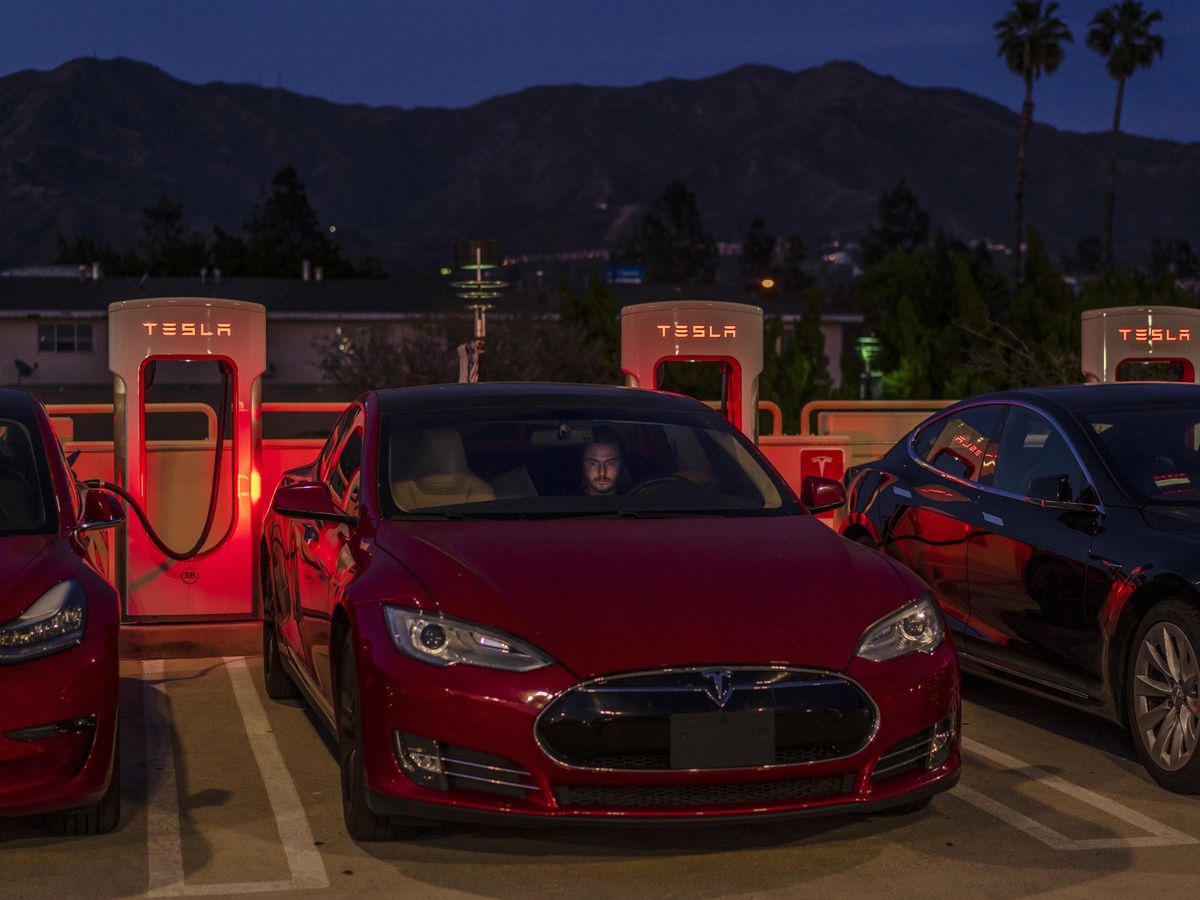 Nighttime photograph of a man in a car at an outdoor Tesla charging lot.
