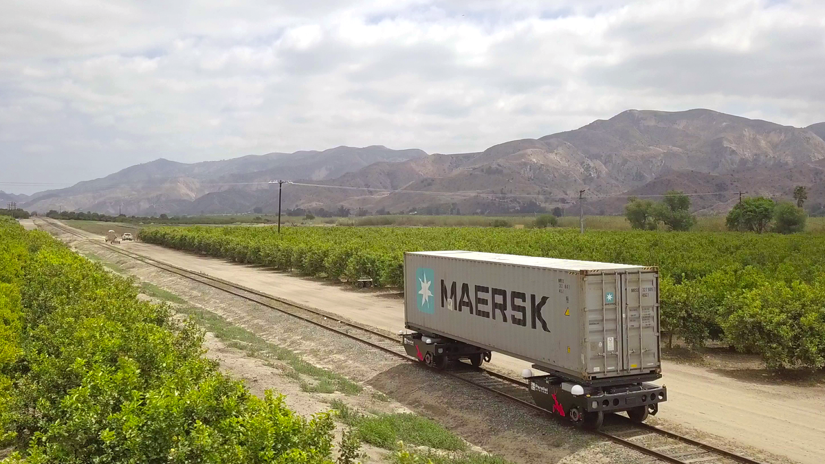 On an isolated stretch of railroad tracks, a freight container is supported at both ends by two autonomous four-wheeled rail vehicles